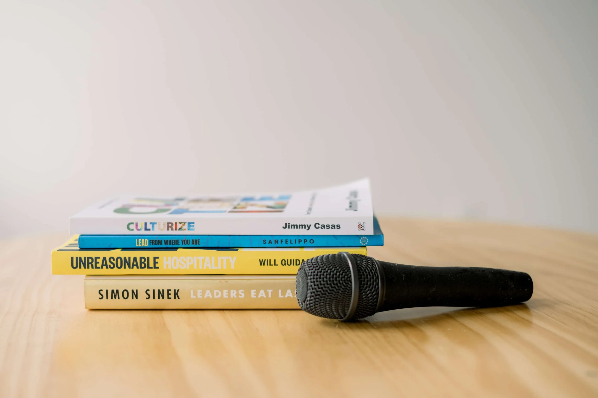 Stacked books on a wooden table with a microphone in front of them, against a plain light-colored background.