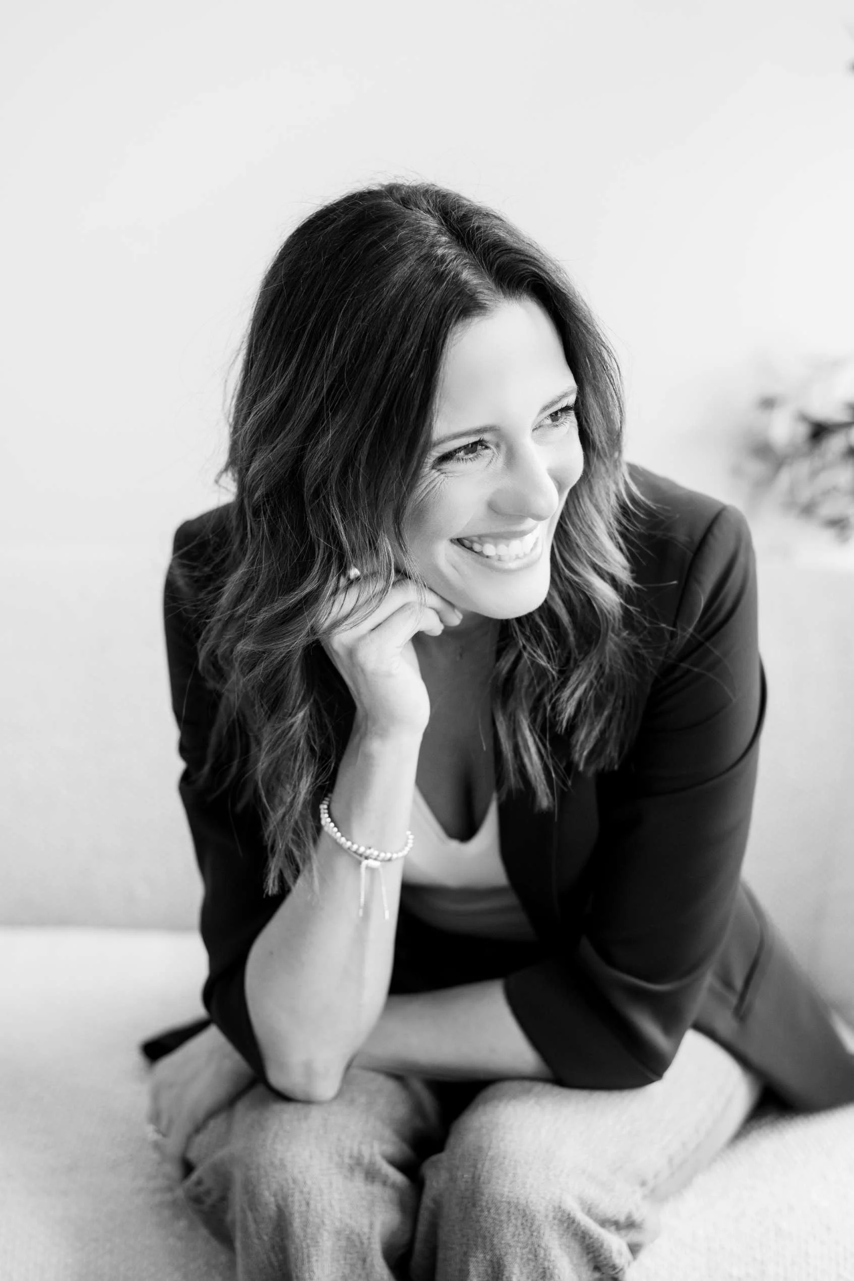 A woman with medium-length wavy hair, smiling and sitting cross-legged on the floor, wearing a dark blazer and light-colored pants, with her hand resting under her chin.