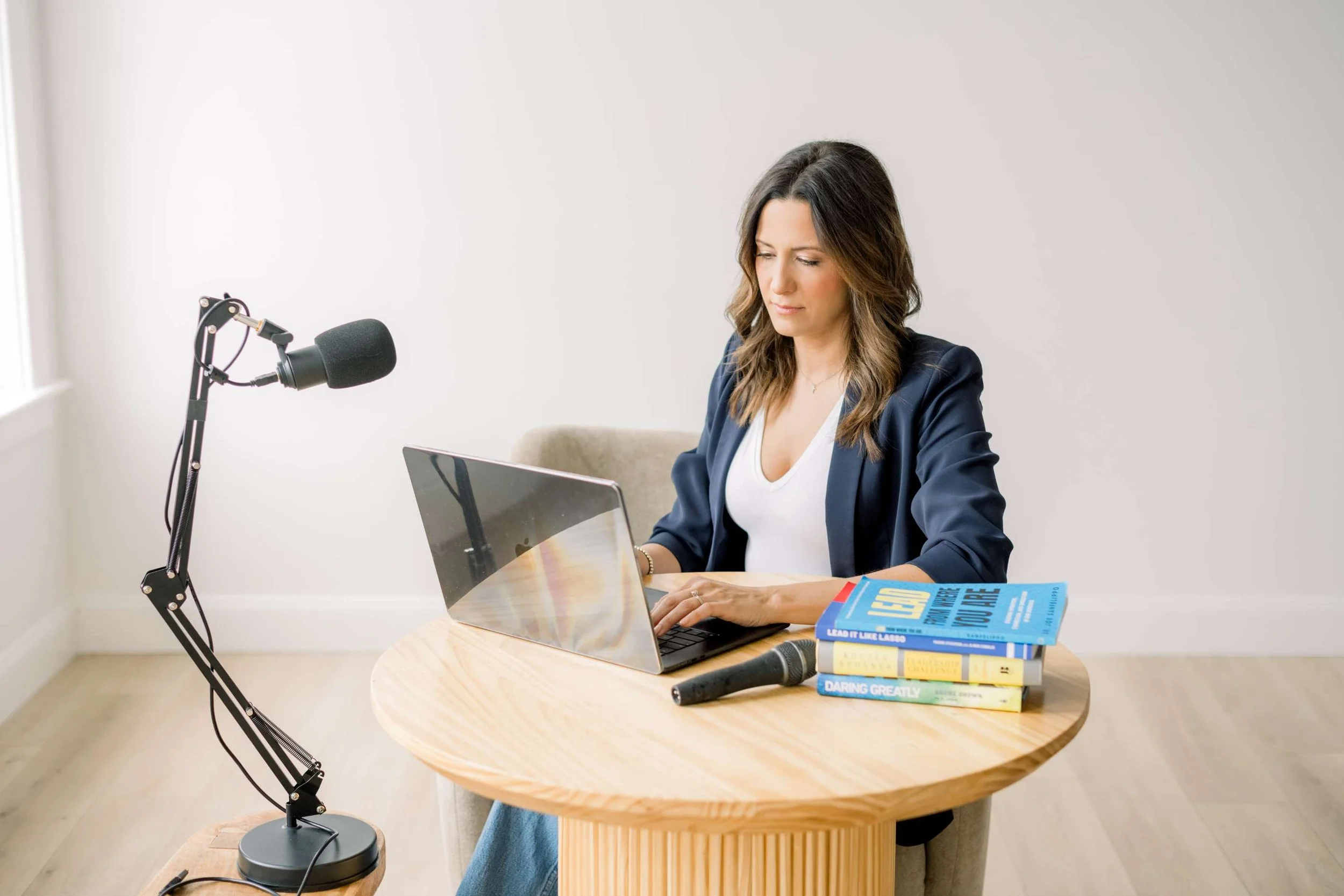 A woman sitting at a round wooden table with a laptop, books, and a microphone, in a minimalist room with white walls.