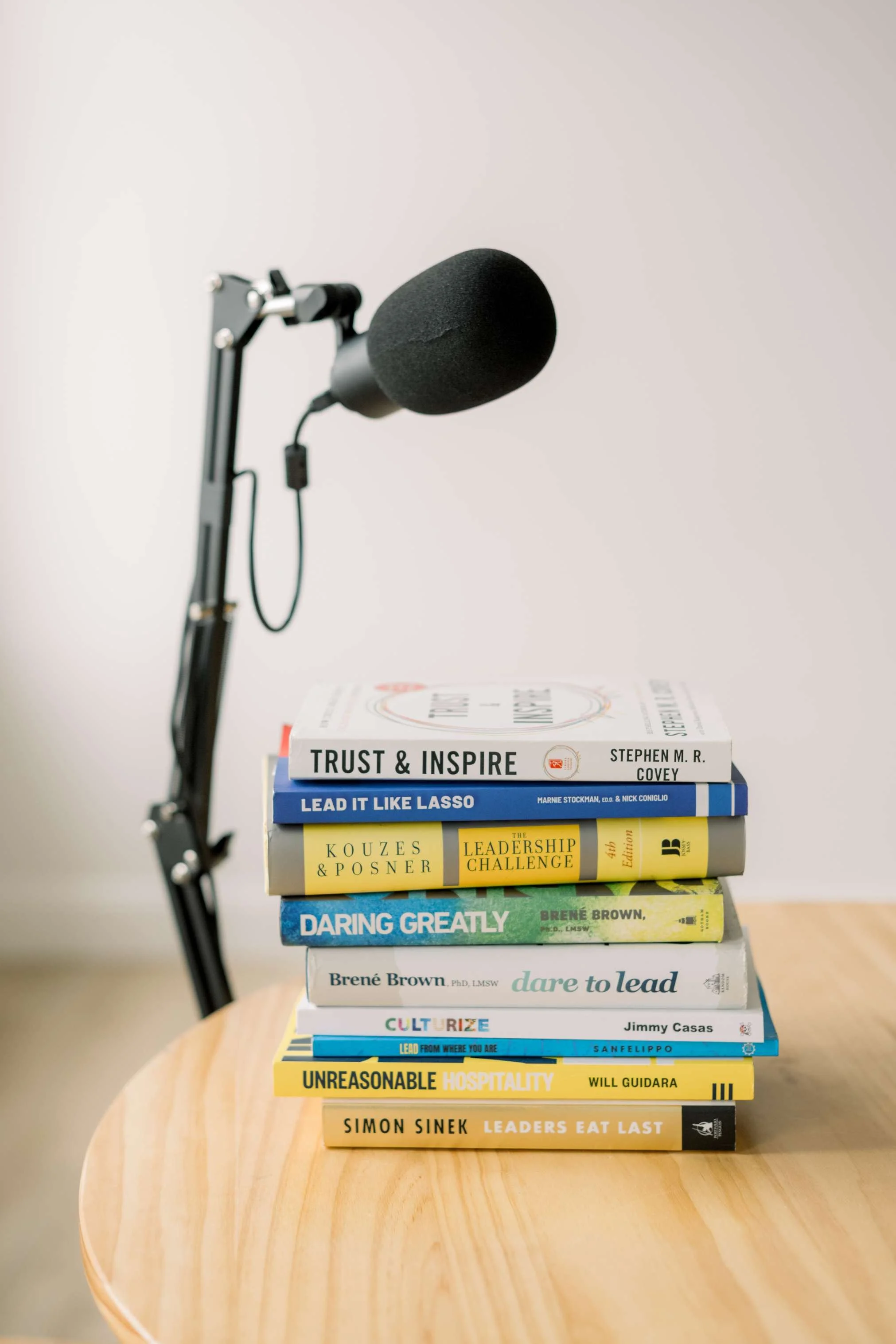A stack of eight books on a light-colored wooden surface with a black microphone on an adjustable arm in the background.