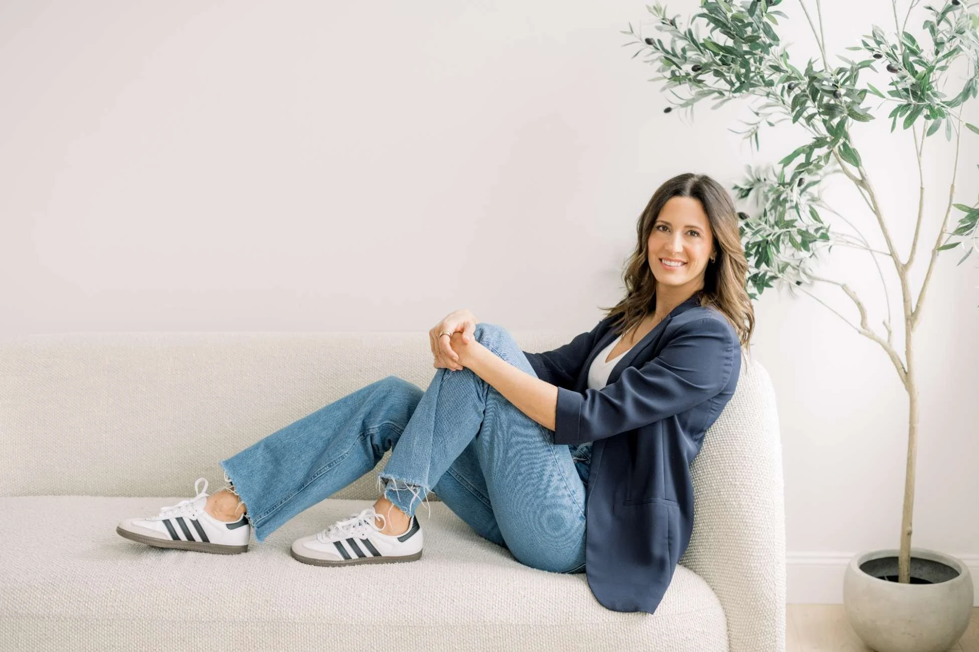 A woman sitting on a beige couch, smiling, with her legs crossed, wearing a navy blazer, white shirt, ripped jeans, and Adidas sneakers. She has shoulder-length brown hair and is positioned next to a potted olive tree in a room with white walls.