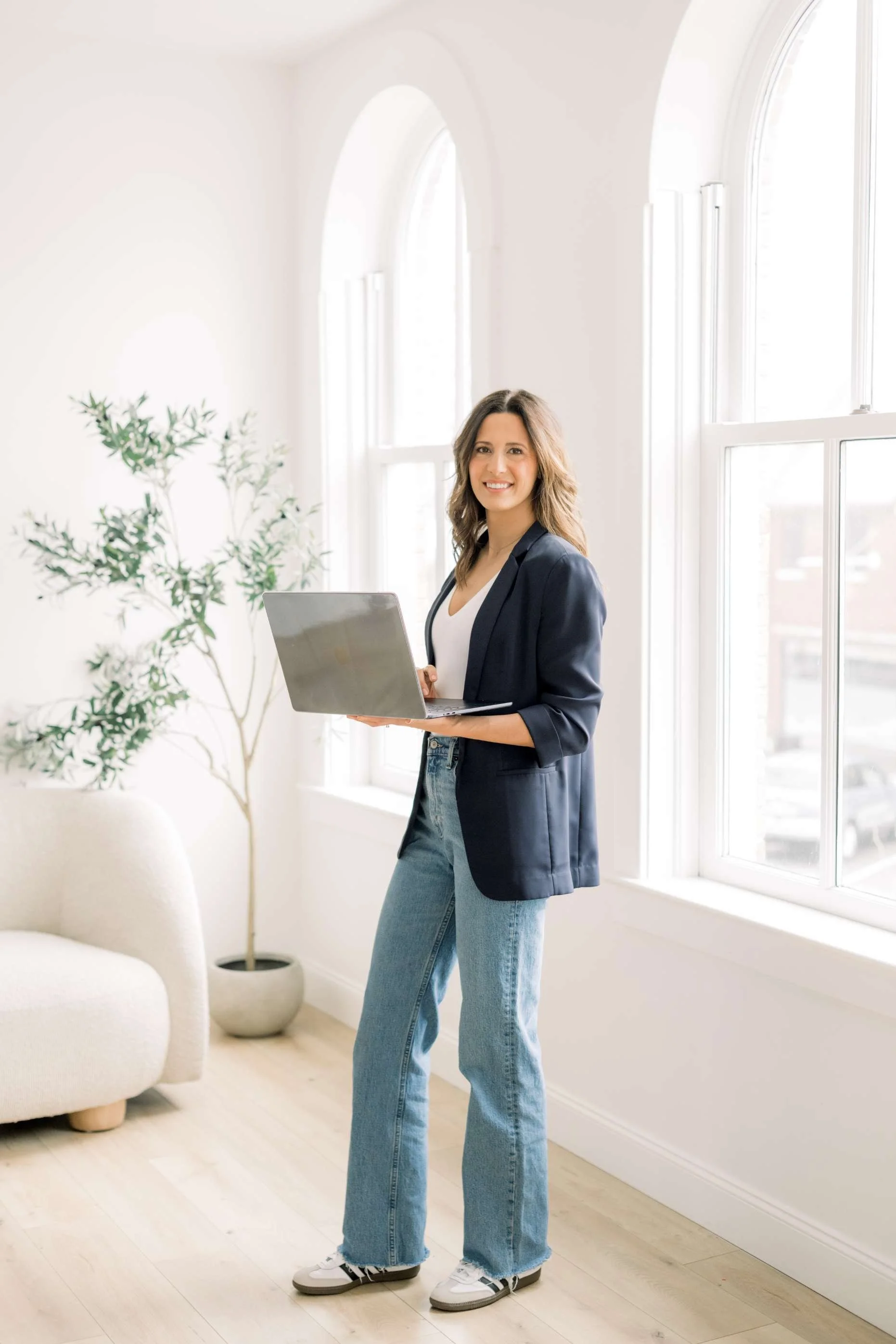 A woman standing indoors near large windows, smiling, holding a laptop, dressed in a blazer, white top, and jeans.