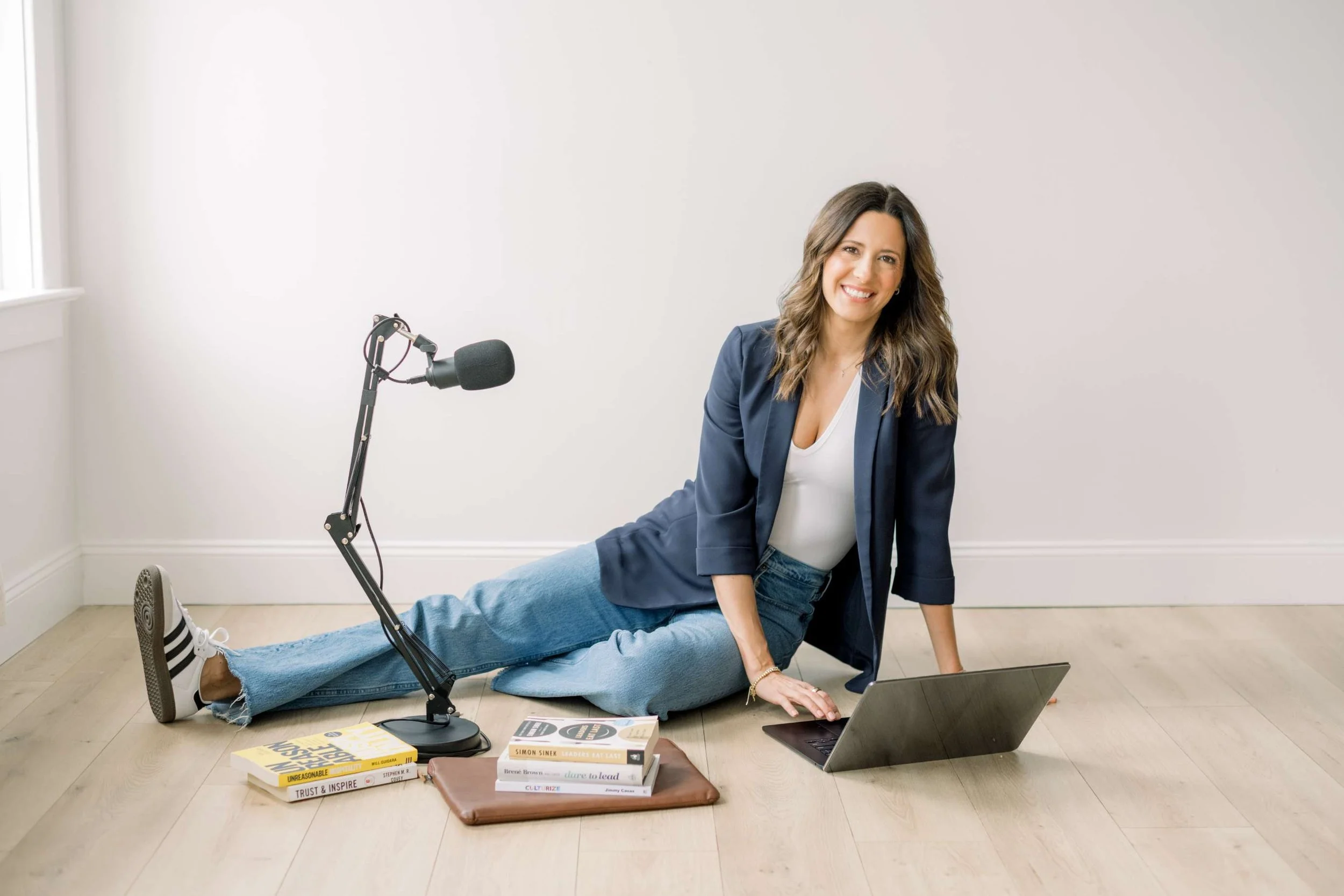 A woman with long wavy hair, smiling, is sitting on the floor in a bright room. She is dressed in a navy blazer, white top, and jeans. She is in a split pose with one leg extended forward and one arm resting on her knee. There is a microphone on a stand, open laptop, and books on the floor nearby.