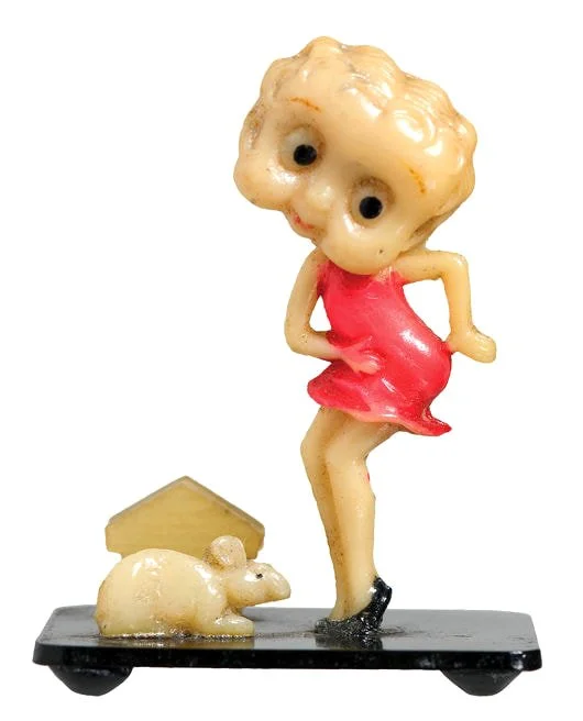 1930s Card Holder Betty is made of celluloid and is 2 inches tall. She stands on a black 2 x ½ inch Bakelite base. A ½ inch celluloid rat or rabbit affixed to the base, along with a celluloid backing to hold a business card between the two. The manuf