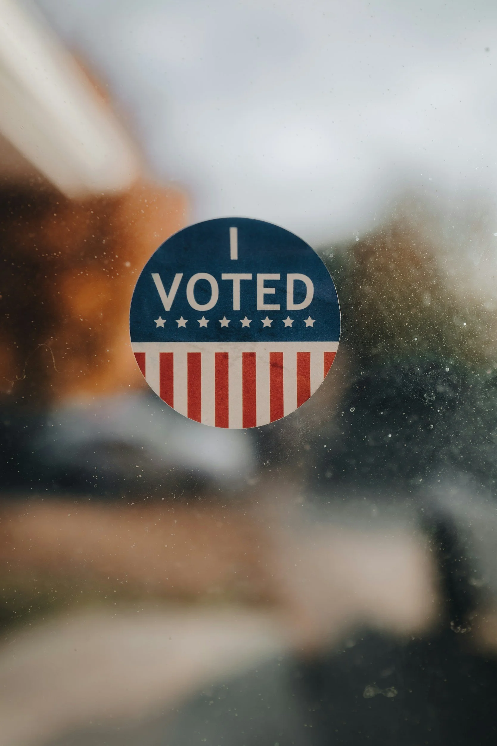 A circular sticker on a window with the text 'I VOTED' in blue and white, with red and white stripes at the bottom.