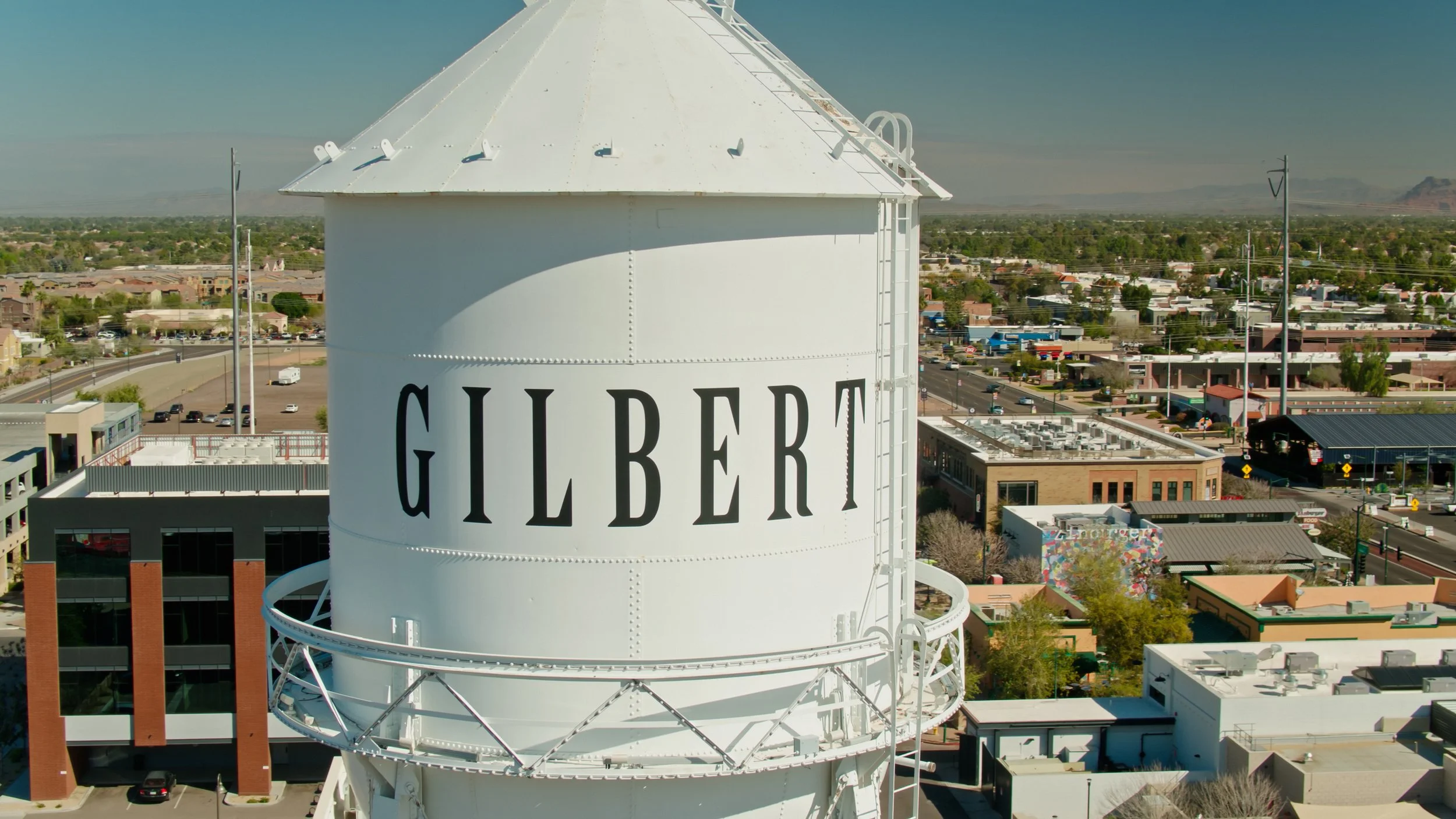 A large white water tower with the word GILBERT written in black, cityscape and suburban area in the background under a clear sky.