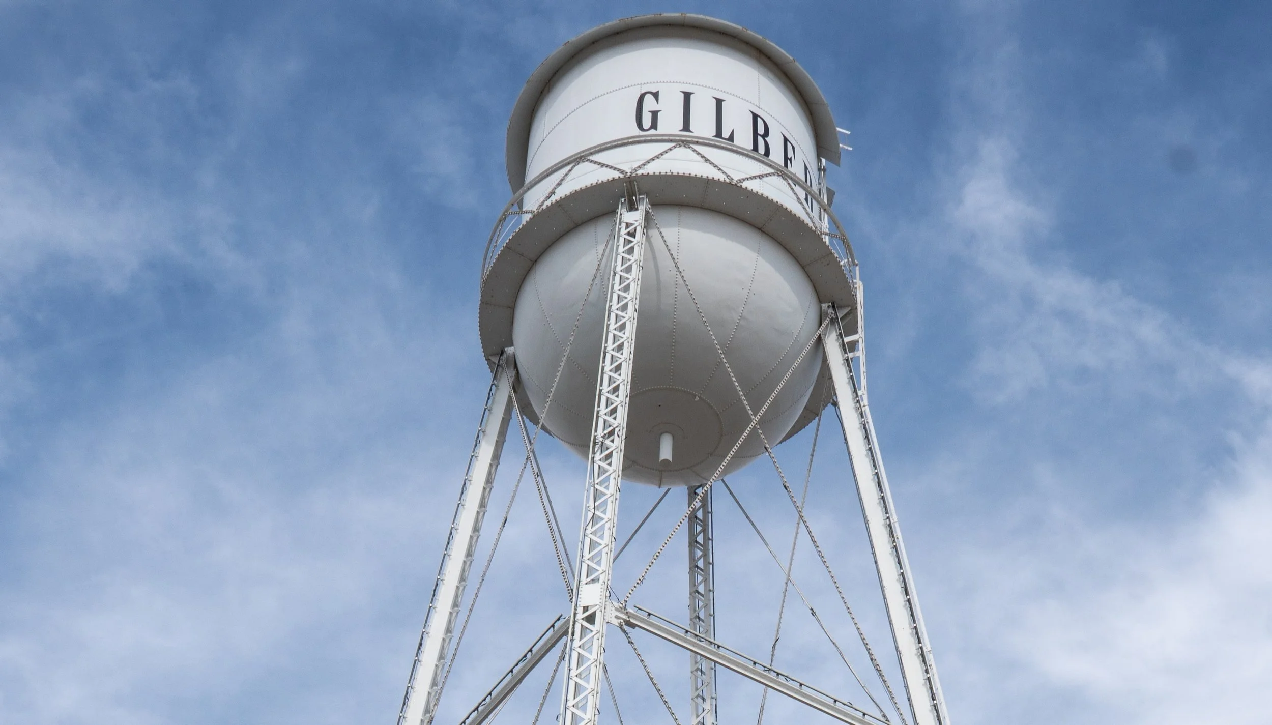 A white water tower with the word 'GILBE' visible on the top portion, supported by metal framework, against a blue sky with some clouds.