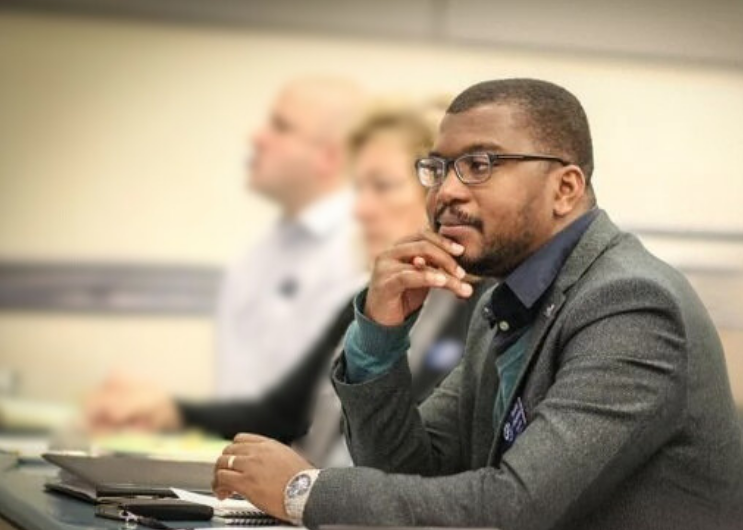 Man in business attire wearing glasses, sitting at a conference table, and listening attentively in a meeting or seminar.