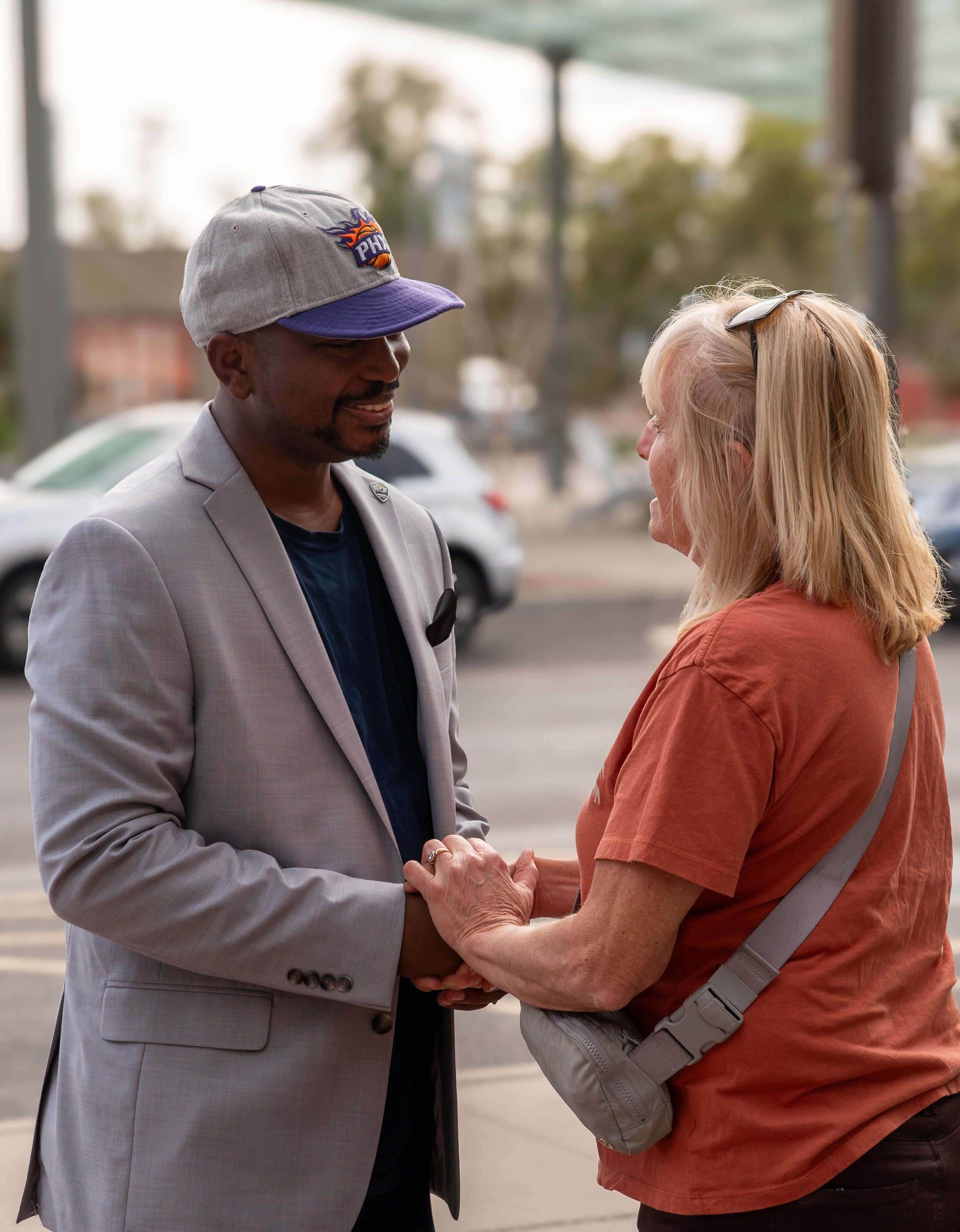 A man and woman smiling and holding hands outside, with cars in the background.