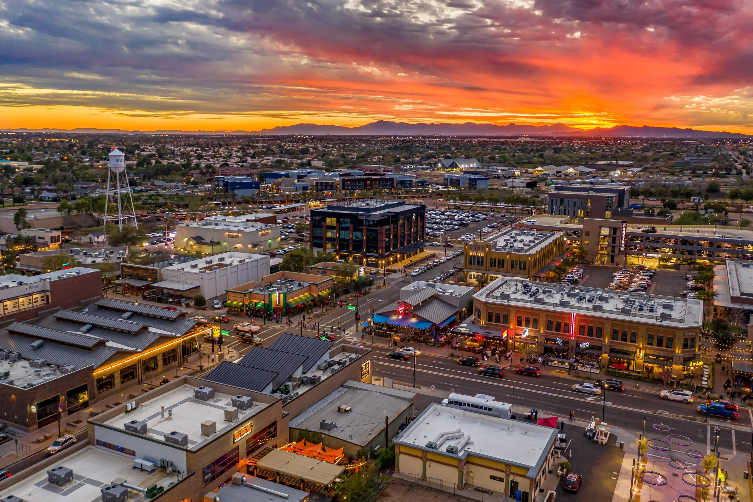A vibrant cityscape during sunset featuring various commercial buildings, restaurants, parking lots, and a water tower with mountains in the background.