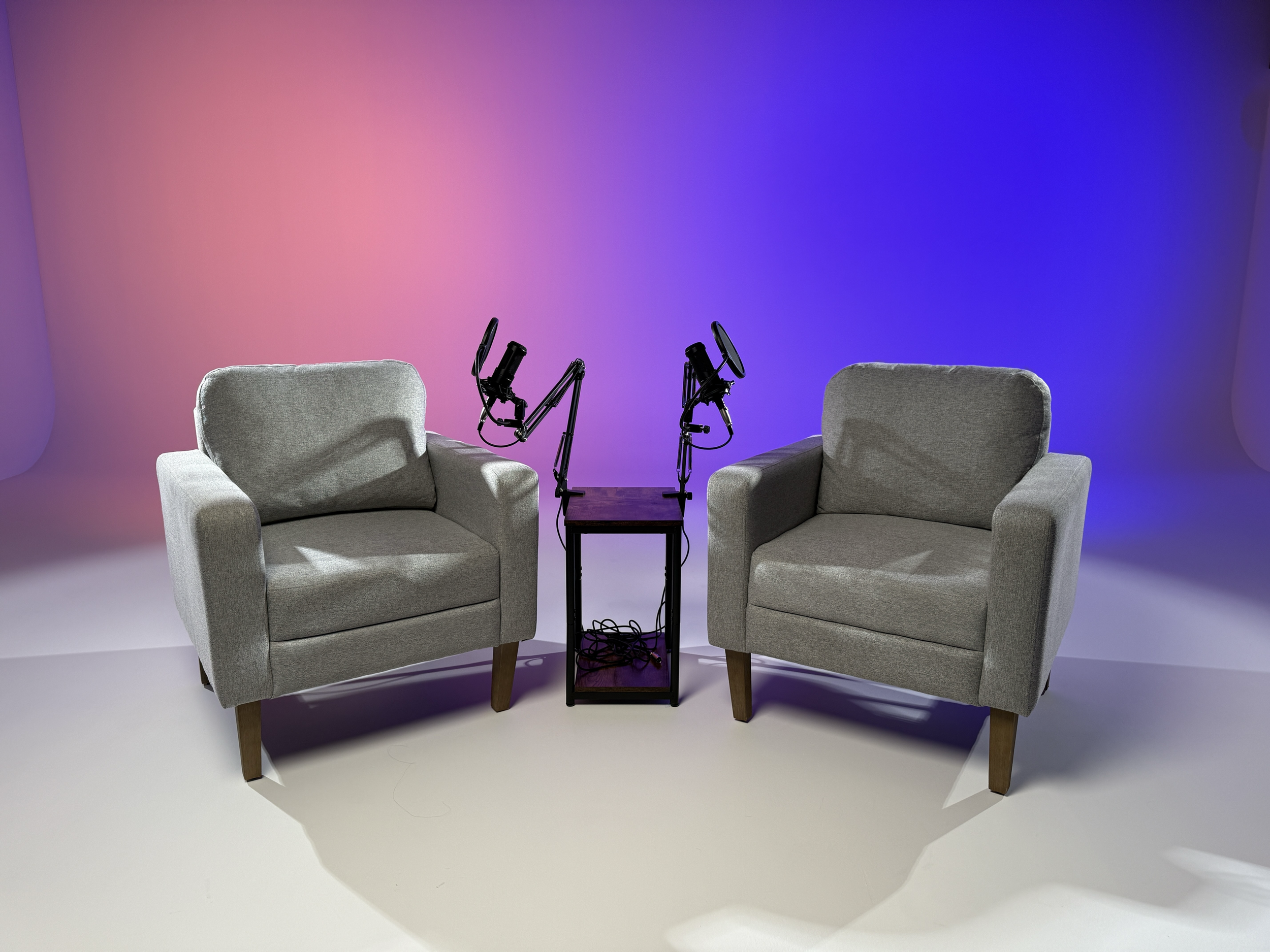 Two gray armchairs facing each other with a small table in between. On the table are two camera mounts or robotic arms. The background has a gradient lighting with pink and blue hues.