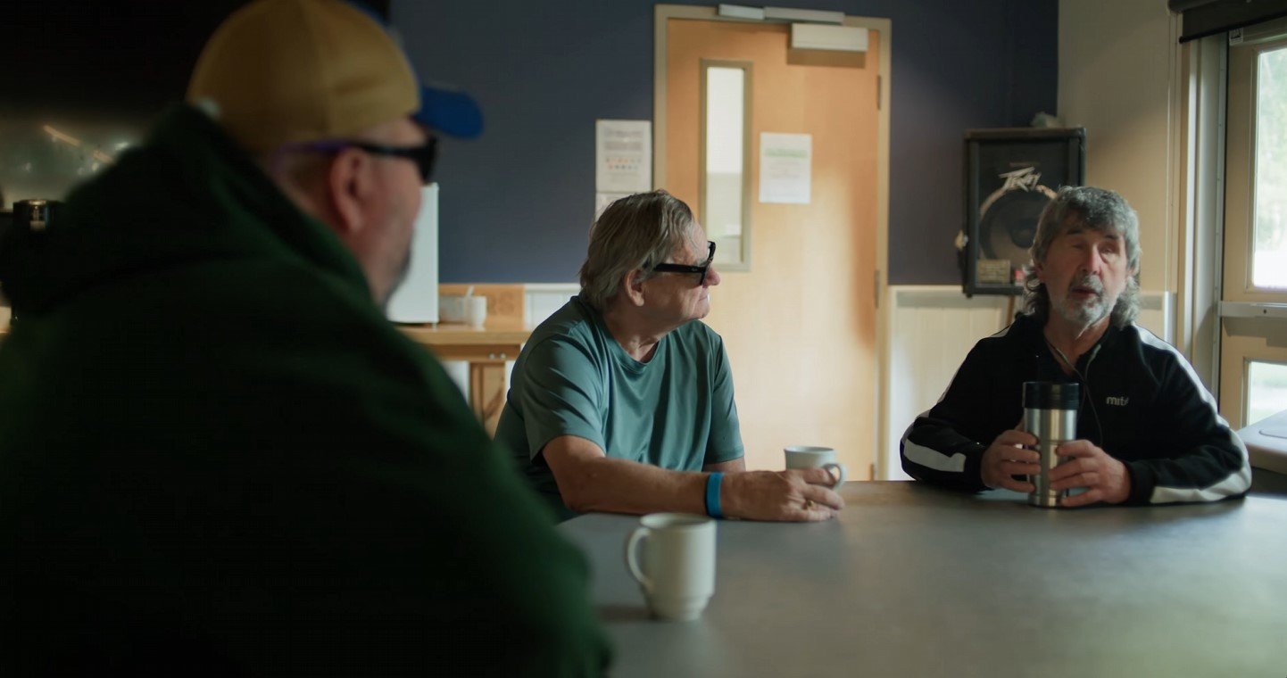 Three men sitting at a table in a room having a conversation, each holding a mug or tumbler.