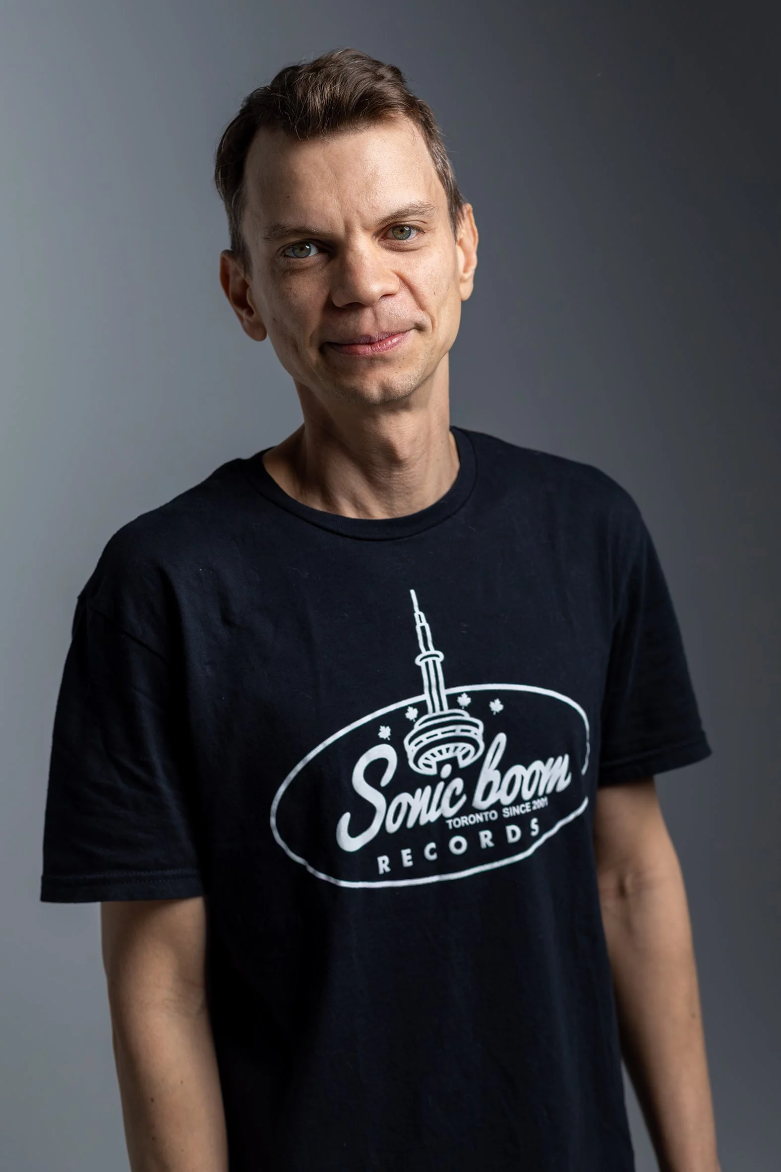 A man wearing a black T-shirt with a logo that says "Sonic Boom Records" and features an illustration of the CN Tower, standing against a plain gray background.