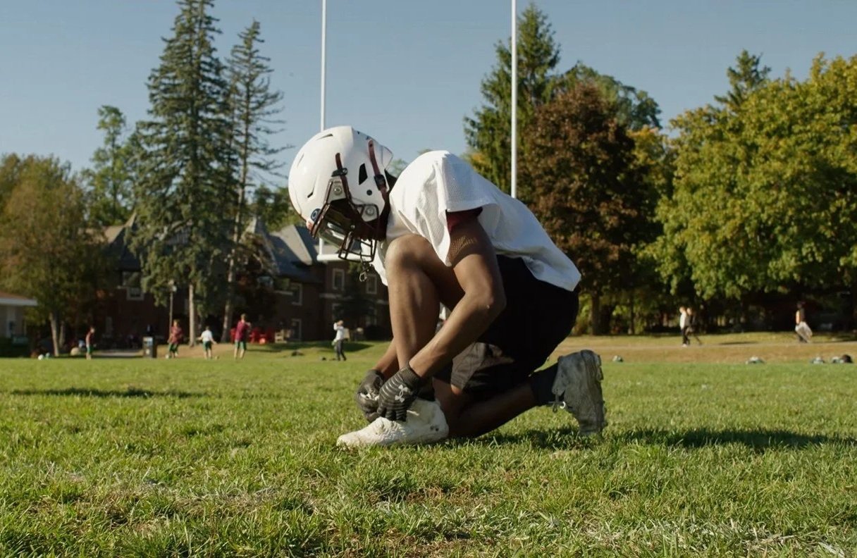 A football player kneeling on a grassy field, holding his shoe, wearing a helmet and football uniform, with trees and other players in the background.