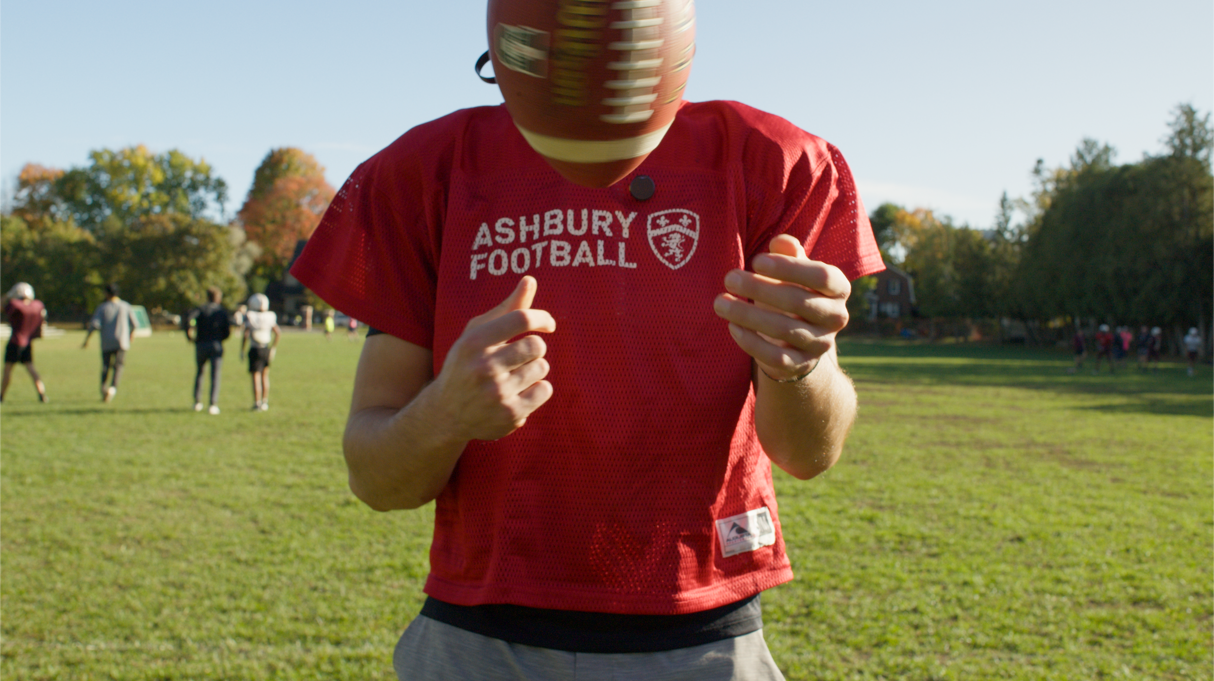 A football player wearing a red Ashbury Football shirt with a helmet, standing on a grassy field with other players in the background and trees with fall foliage.