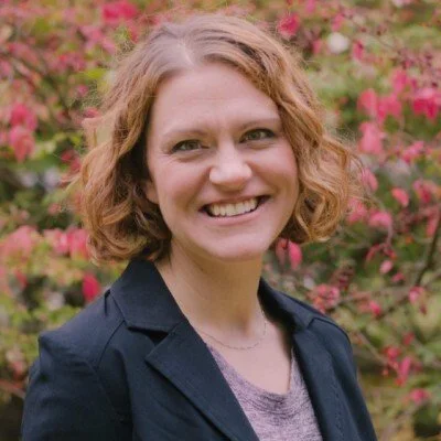 A middle-aged man with gray beard sitting on a bench against a plain wall, wearing a brown polo shirt and cream-colored pants, looking contemplative.