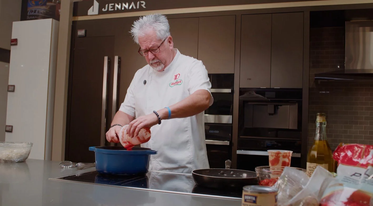 A man with gray hair and glasses, wearing a white chef's coat, is preparing food in a modern kitchen, pouring ingredients into a blue bowl.
