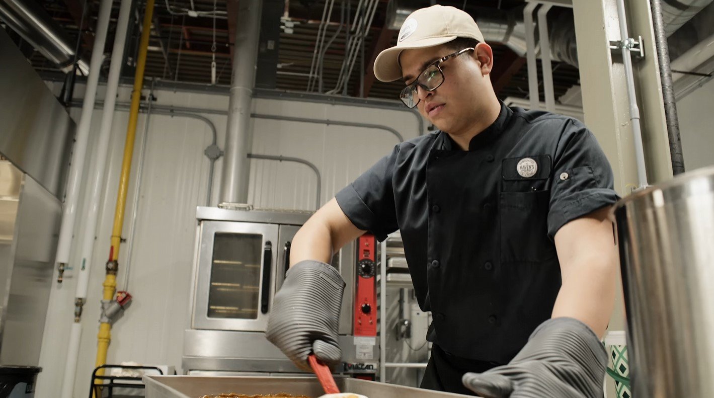 Young man with glasses and a beige cap preparing food in a commercial kitchen, wearing black work clothes and gray oven mitts.