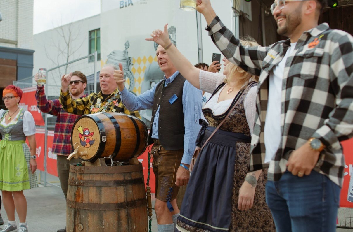 Group of people at an outdoor beer festival, some in traditional German attire, raising glasses and giving thumbs up, with a wooden beer barrel and a cartoon beer tap logo in the foreground.