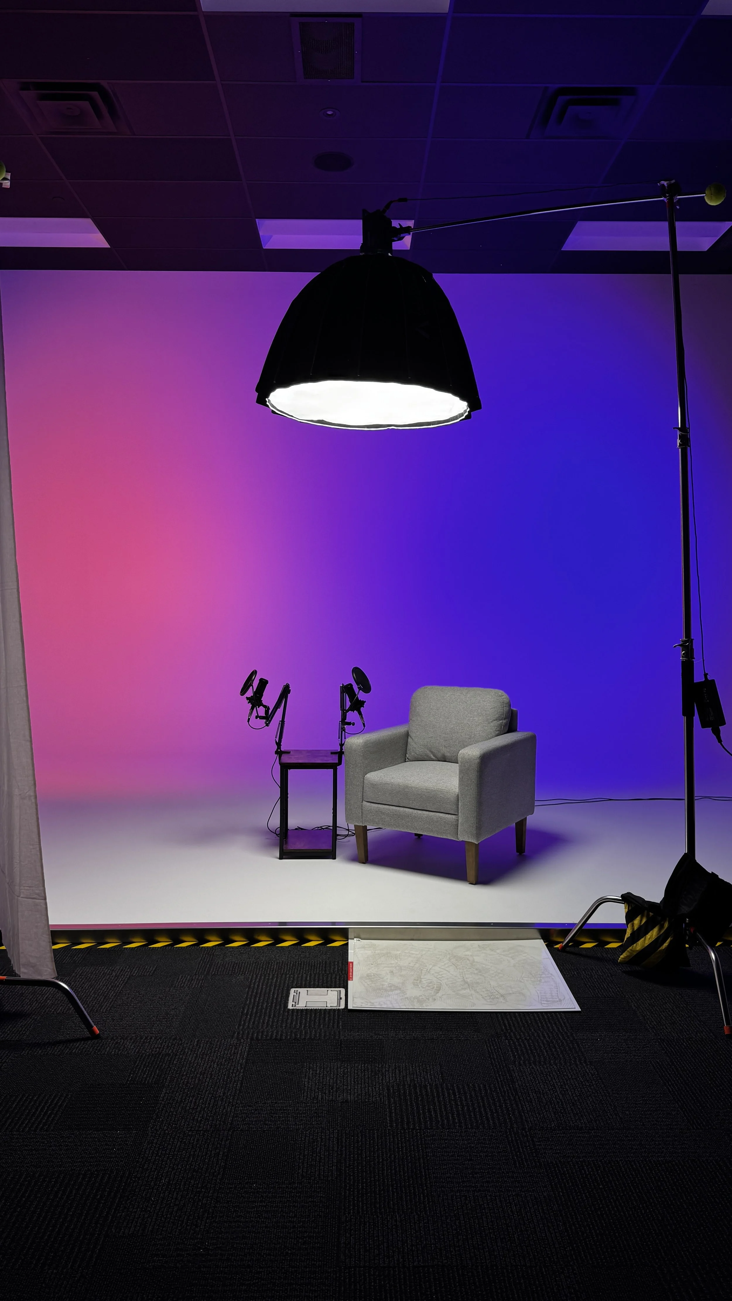 A photography studio setup with a gray armchair, professional lighting equipment, and a backdrop with purple and pink gradient lighting.