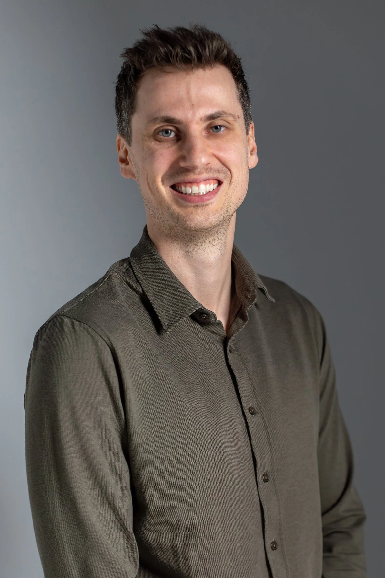 A man smiling, wearing a dark gray button-up shirt, standing against a gradient gray background.