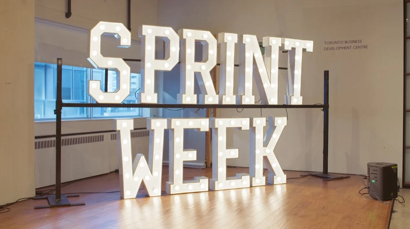 Large white illuminated sign reading 'SPRINT WEEK' in a room with hardwood floors and windows.