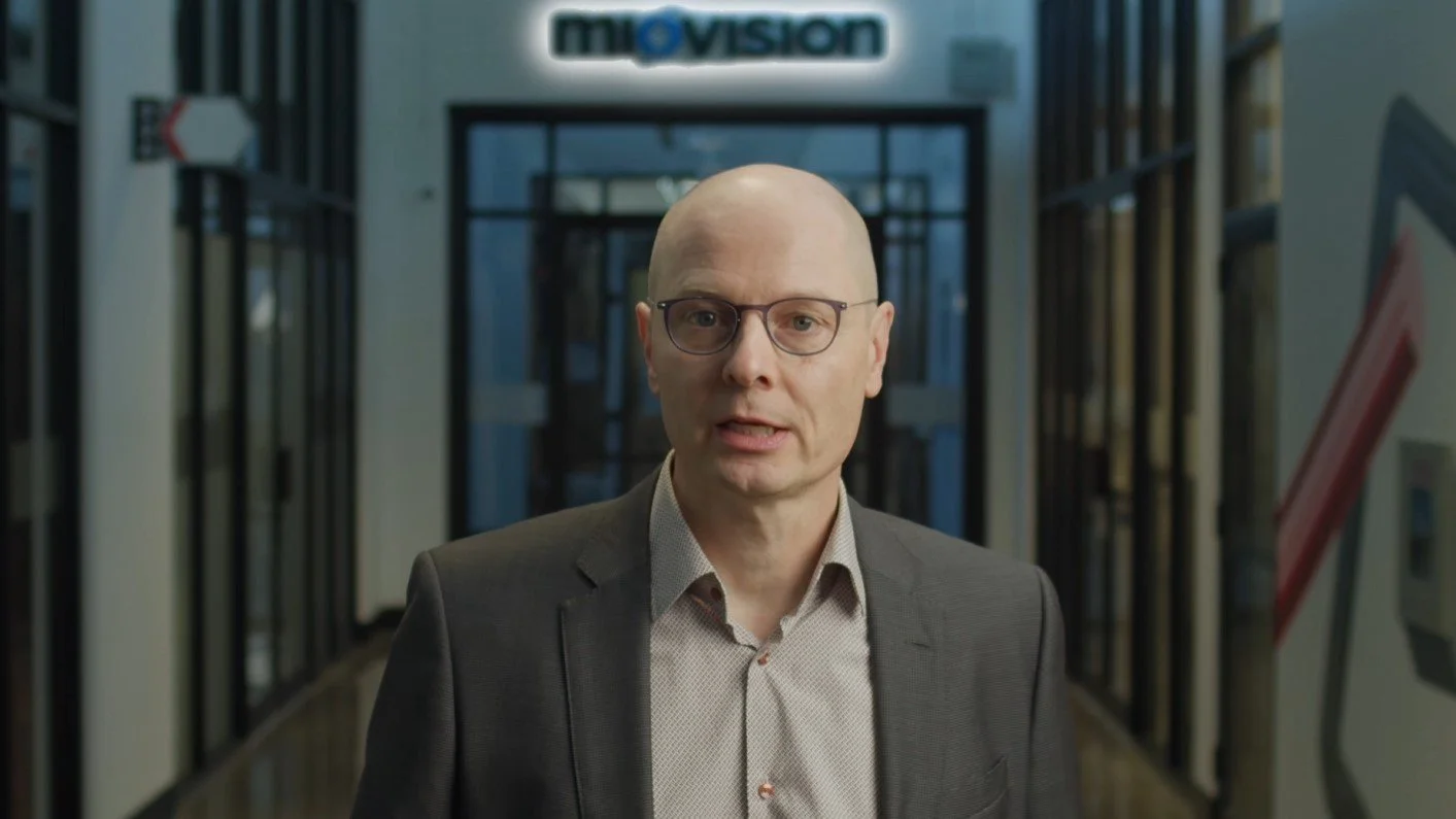 A bald man with glasses wearing a gray suit and light-colored shirt standing in front of a sign that says 'movision' inside a building.