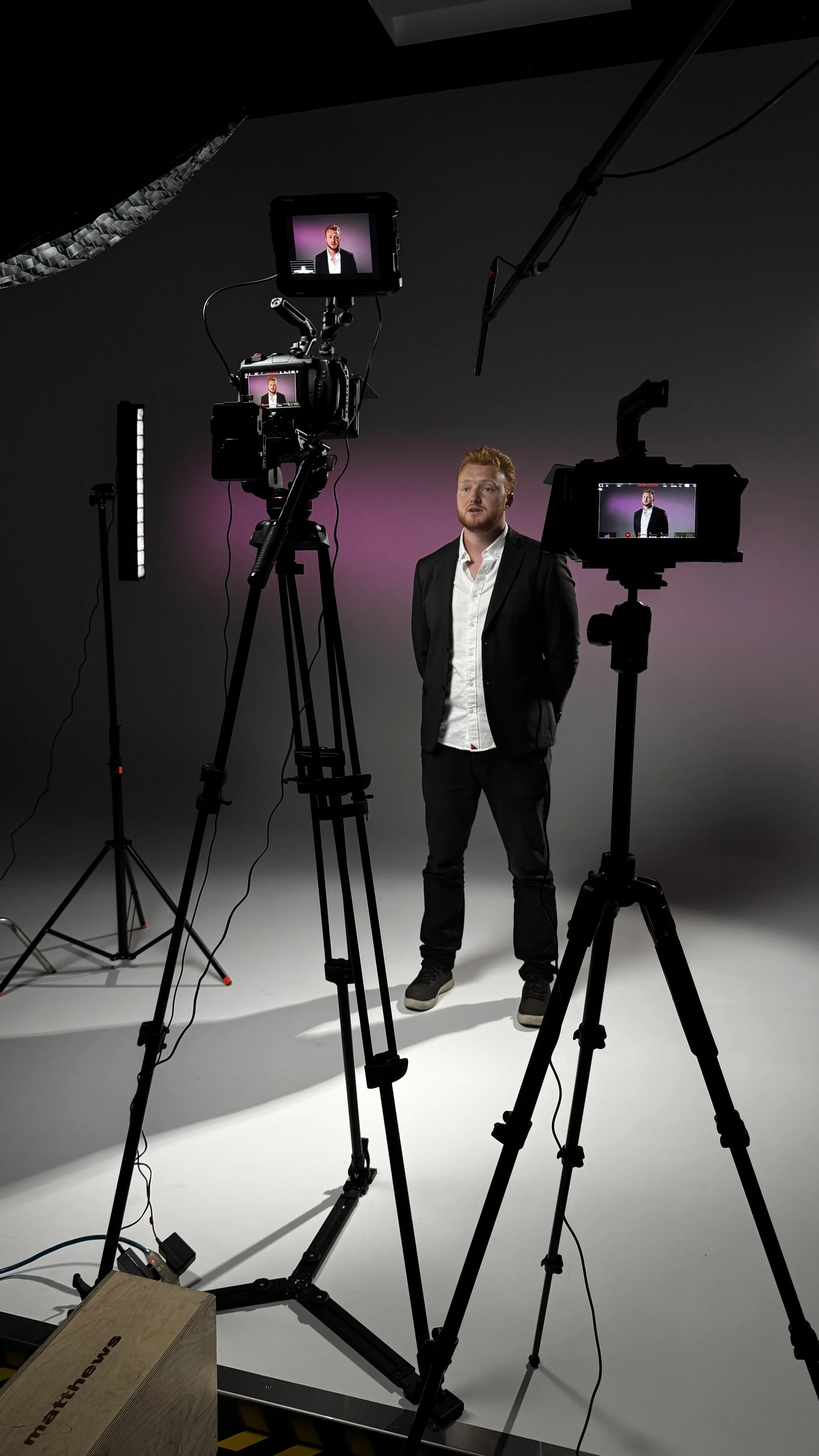 A man in a black suit and white shirt standing in a professional video recording studio with cameras and lighting equipment, filmed from multiple angles.