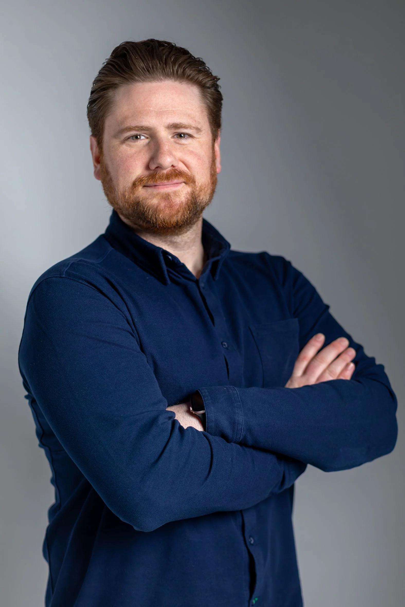 A man with red hair and beard wearing a dark blue button-up shirt, standing with arms crossed against a plain gray background.