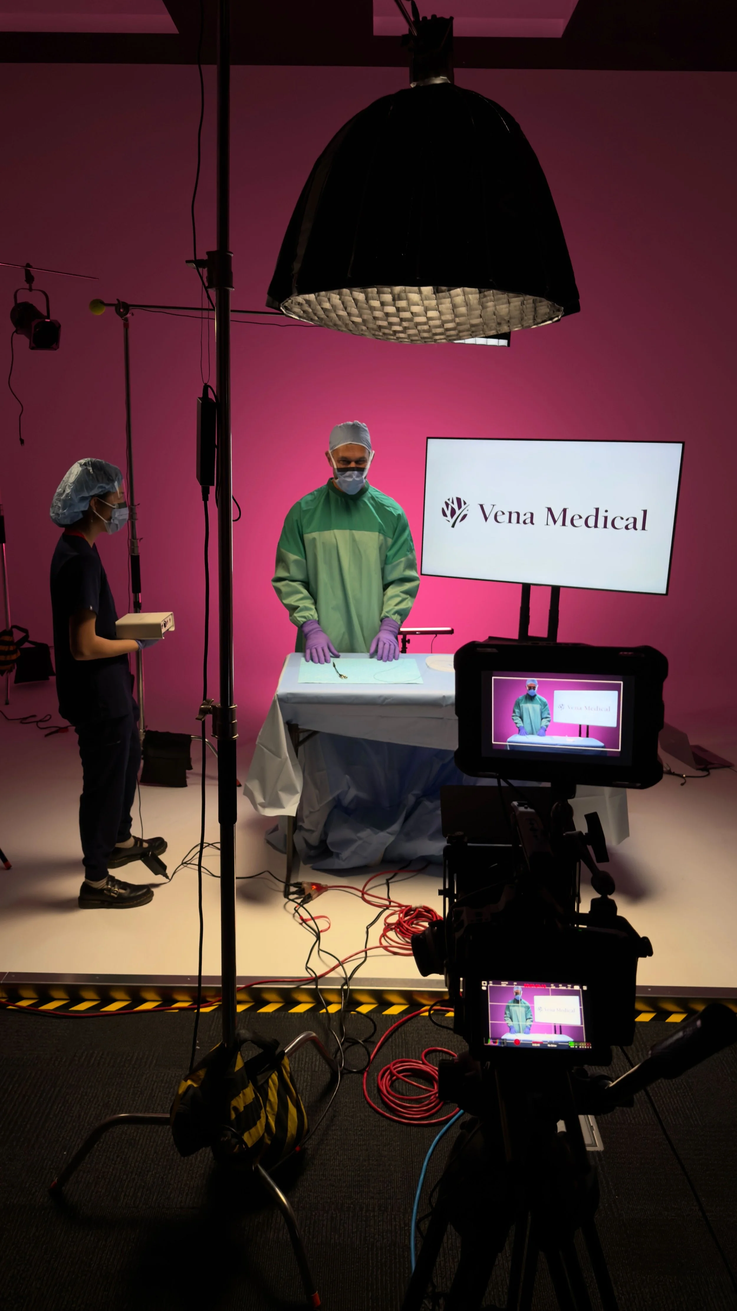 A medical professional in green scrubs and purple gloves standing behind a table, with a monitor displaying the Vena Medical logo in the background. Two other people, also in medical attire and masks, are behind him in a studio setup with professional lighting and filming equipment.