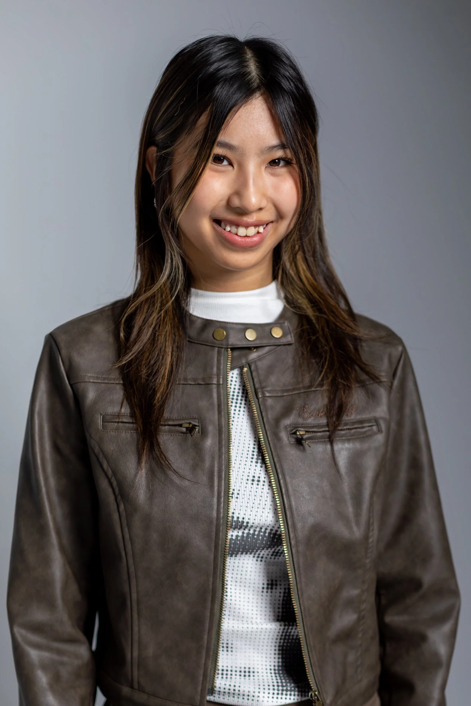 A young woman with dark hair and light skin, smiling, wearing a brown leather jacket over a white top, against a gray background.