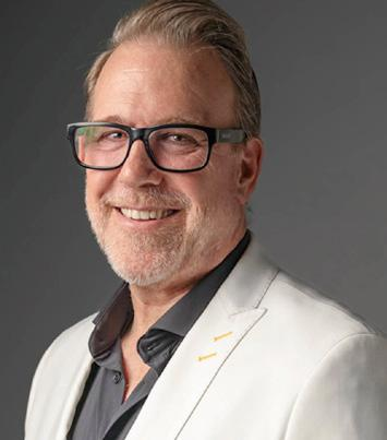 A middle-aged man with glasses and a beard, smiling, wearing a white blazer and a dark shirt, against a dark background.
