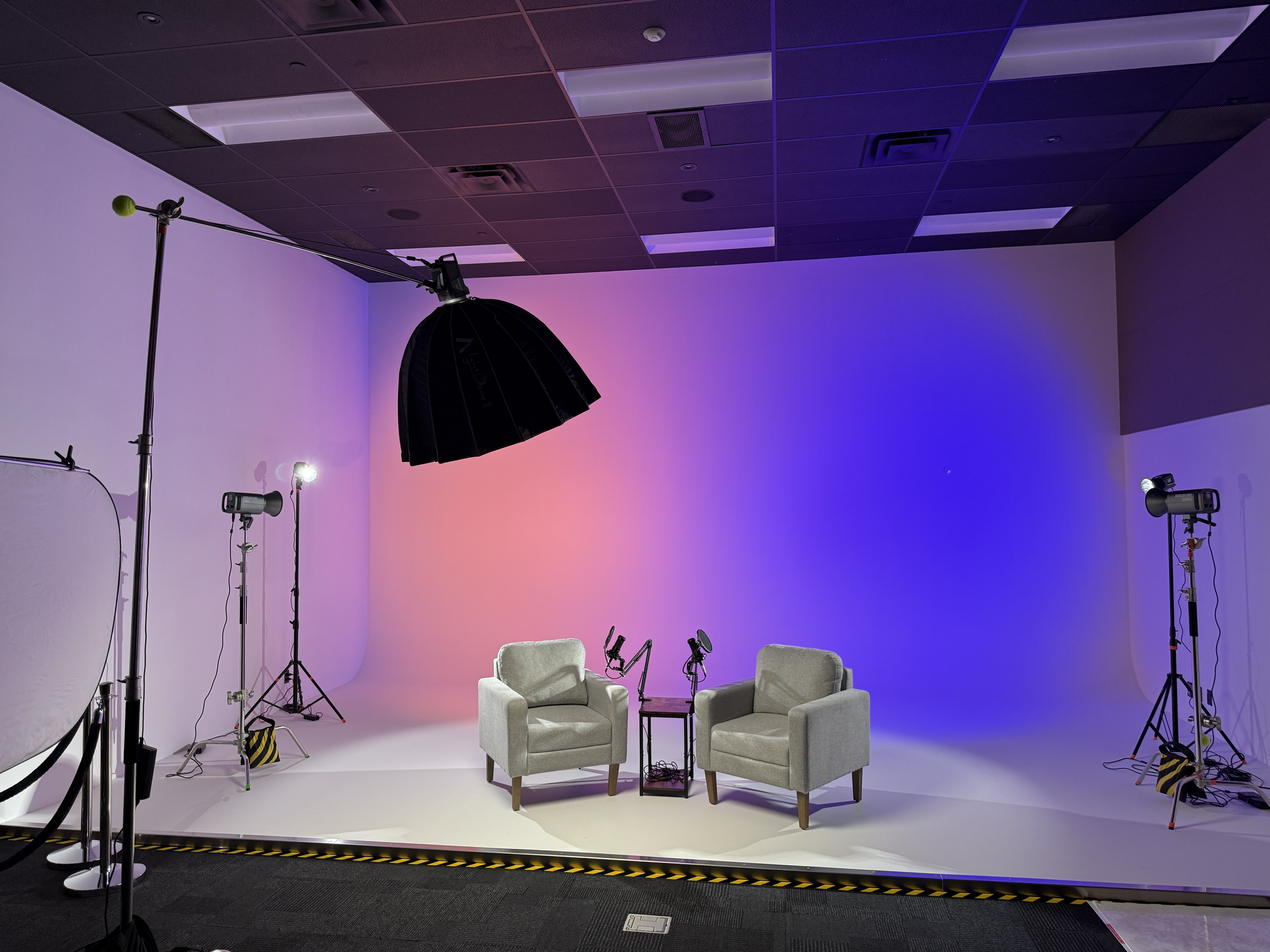 Photo of a photography studio setup with two beige armchairs, a small black table, and photography equipment including lights, umbrellas, and backdrops with a gradient pink and blue background.