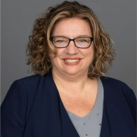 A woman with short curly hair wearing glasses, a navy blazer, and a light blue top, smiling at the camera.