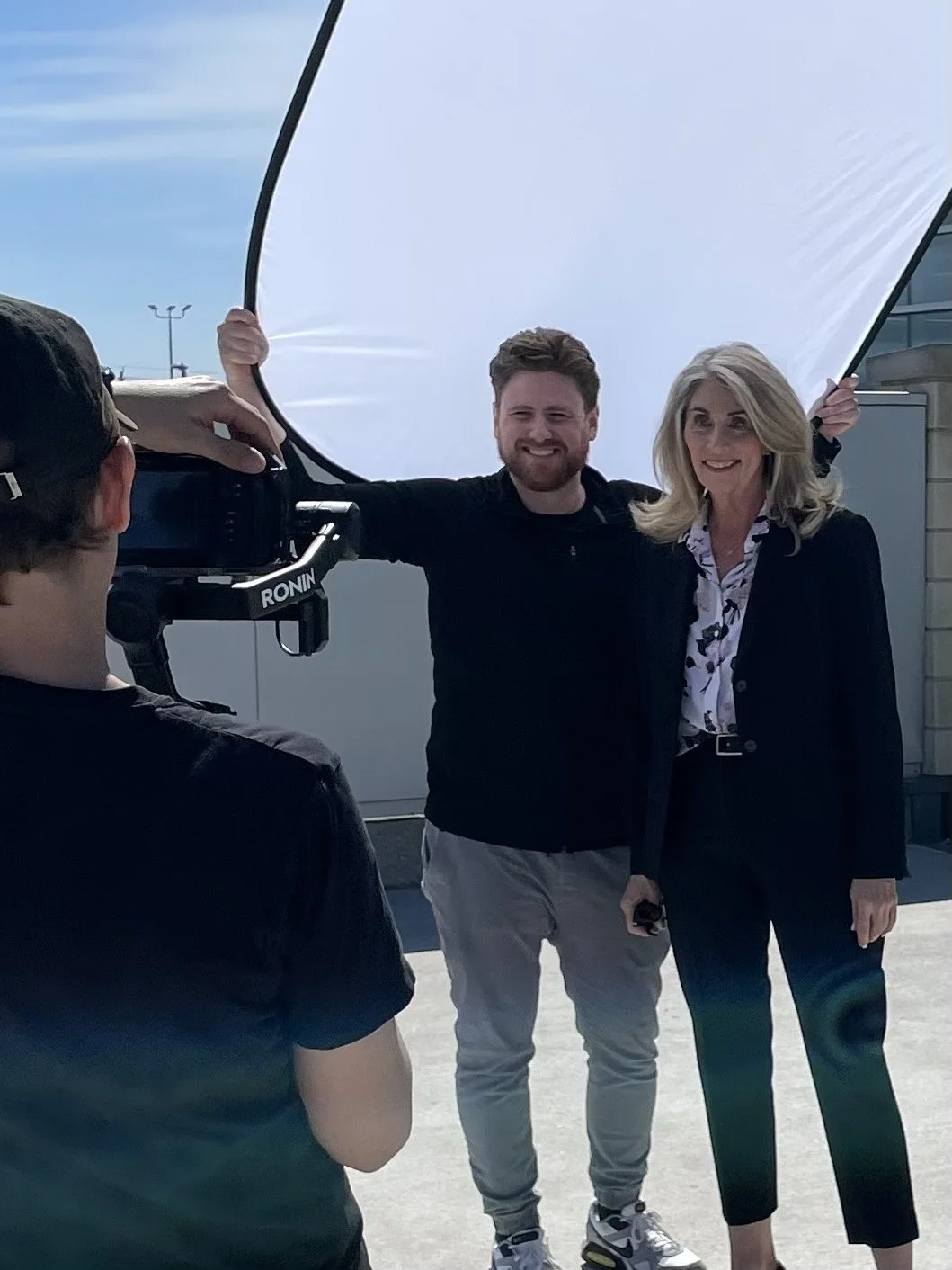 Man and woman pose for a photo being taken by a photographer, with a professional reflector in front of them on a rooftop or outdoor setting.