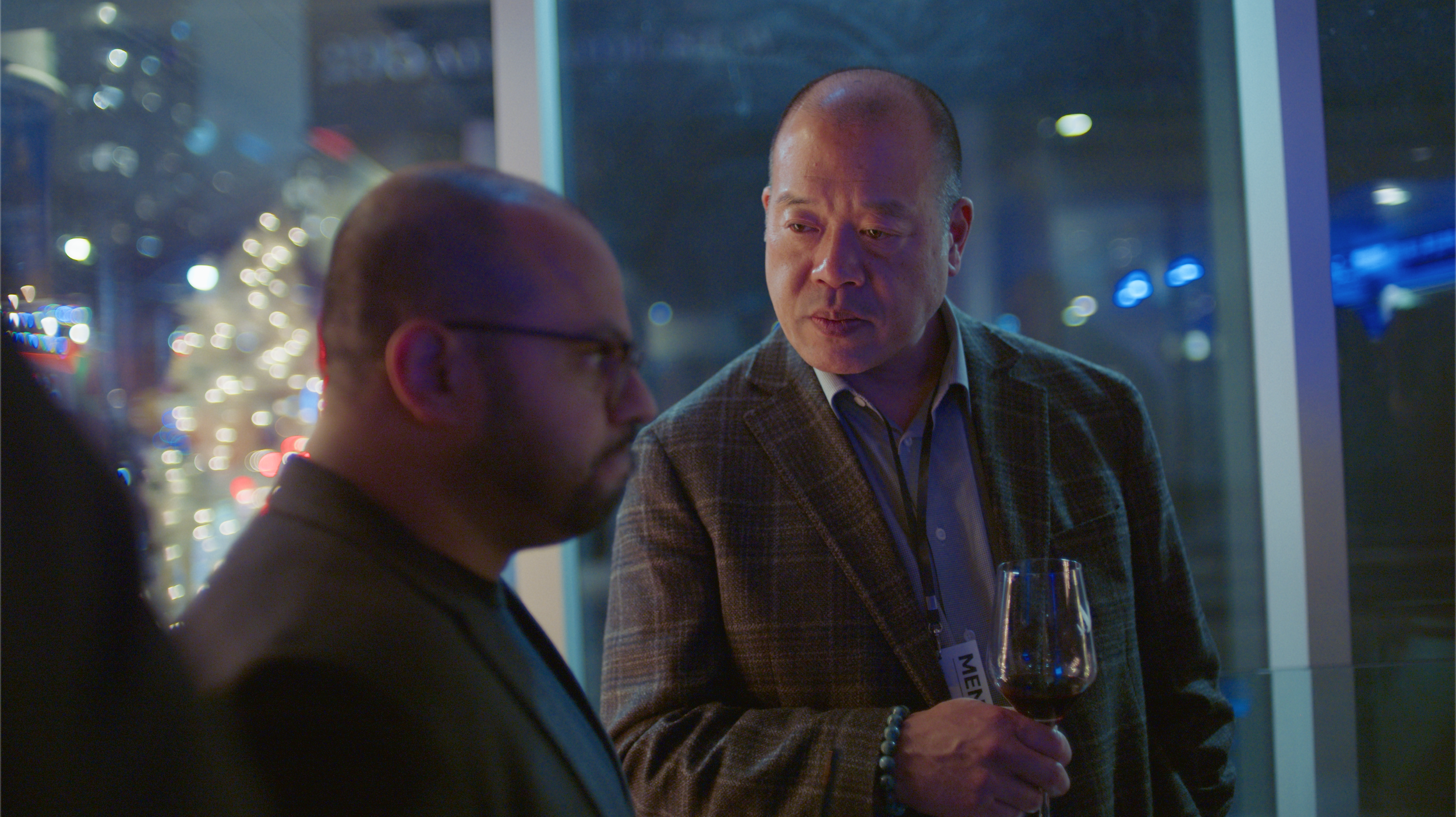 Two men having a conversation at a nighttime event, with city lights visible through the windows behind them. One man is holding a glass of red wine.