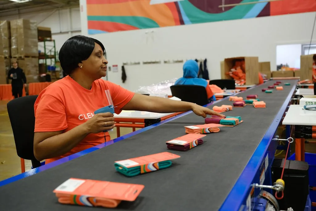 A worker in an orange Cleanlogic t-shirt seated at a long conveyor belt production line, reaching to inspect folded, packaged washcloths as they move down the line.