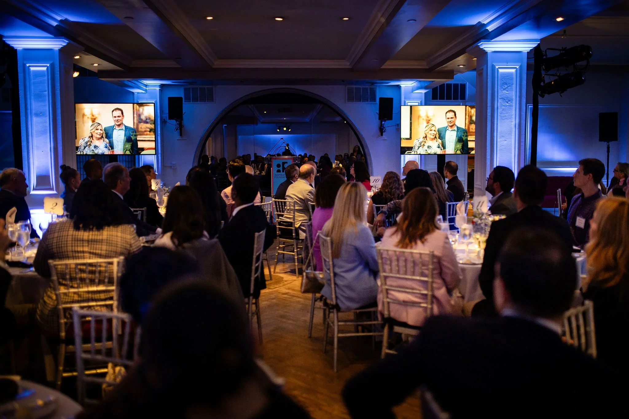 Guests seated at round tables in a ballroom lit with blue uplighting watch a couple on stage displayed on two large screens, with a lectern and "Inspiration Foundation" banner visible in the background.