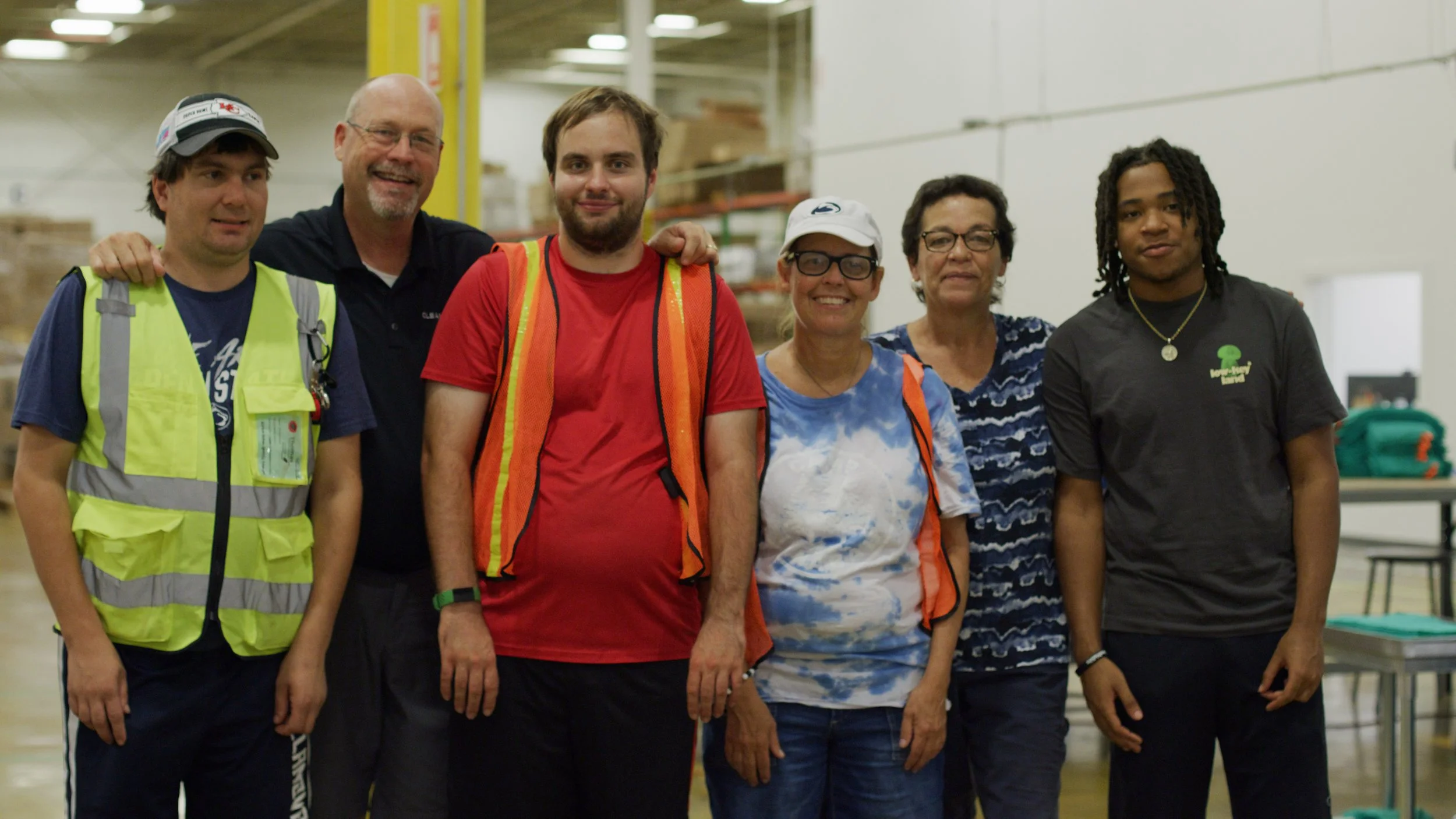 A group of six smiling coworkers pose together in a warehouse, two wearing high-visibility safety vests, standing in front of shelving and storage boxes
