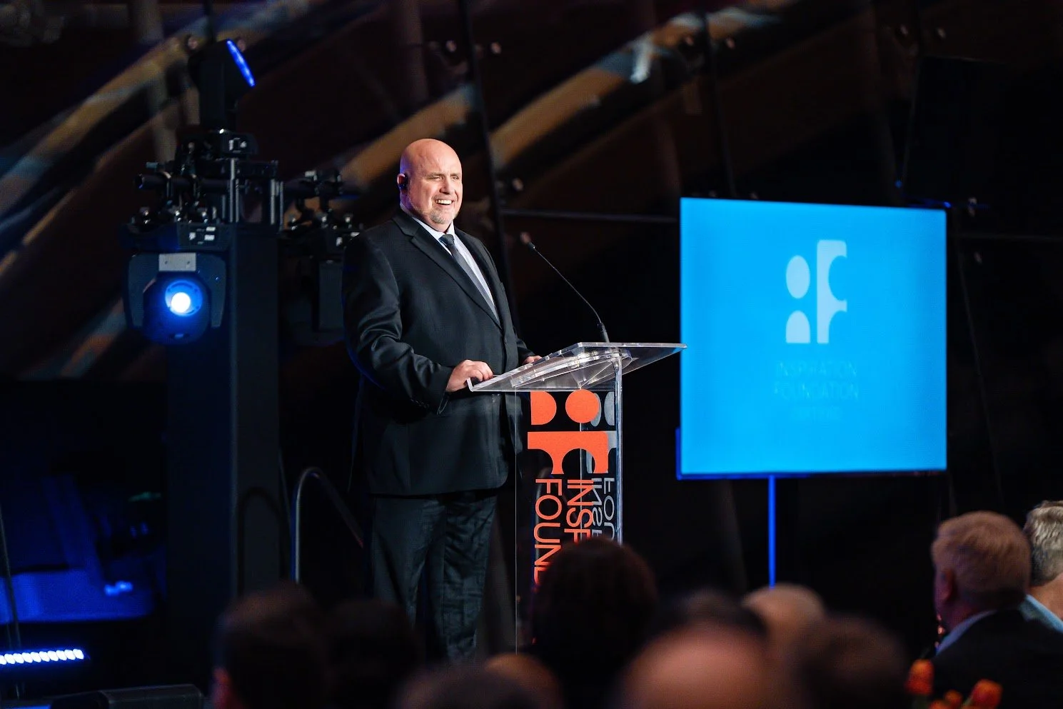 Jeremy Grandstaff is pictured in dark suit he smiles while speaking at a clear acrylic podium branded with the Inspiration Foundation logo, with a large screen displaying the foundation's logo behind him with an audience seated in the foreground