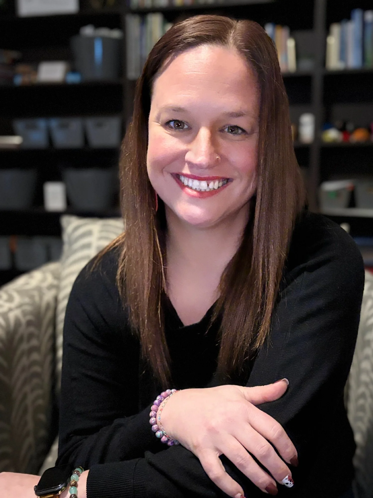 A woman with long brown hair, wearing a black top, smiling, sitting on a patterned chair in a room with black shelves filled with books and small objects behind her.