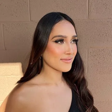 A woman with long, dark hair and makeup, looking to the side against a brick wall background.