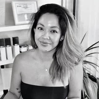 A woman with shoulder-length hair posing and smiling in a room with framed certificates and shelves in the background.