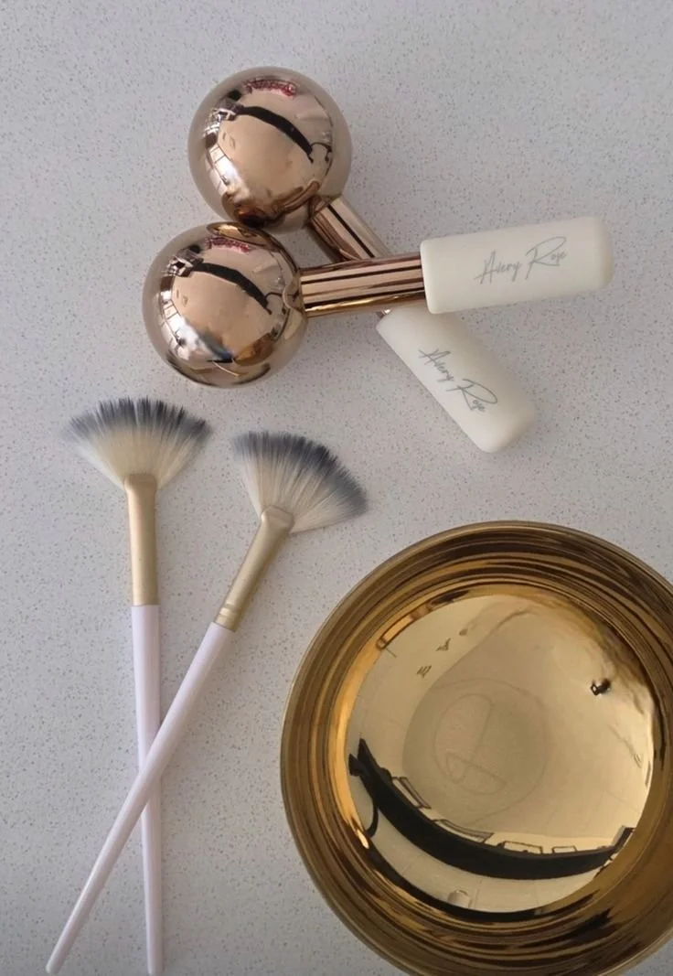 A gold decorative bowl, two fan-shaped cleaning brushes with white handles, a pair of rose gold massage rollers, and two white beauty sponges with the signature 'Harry Rose' on them on a white surface.