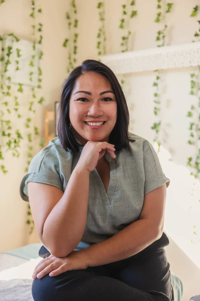 A smiling woman with short dark hair, wearing a light gray top, resting her chin on her hand, seated indoors with a background decorated with green leafy plants and beige walls.