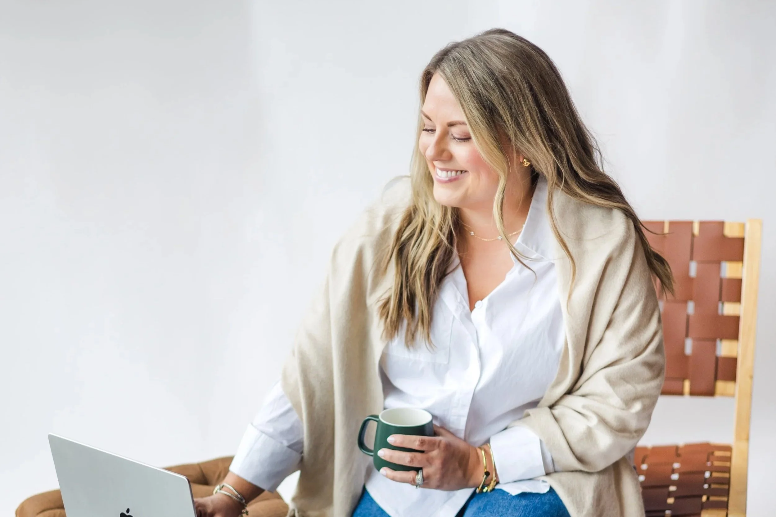 A woman with long, light brown hair sitting at a table, smiling, holding a green mug, with a silver laptop in front of her, wearing a white shirt and beige cardigan. There is a brown chair with a wooden back in the background.