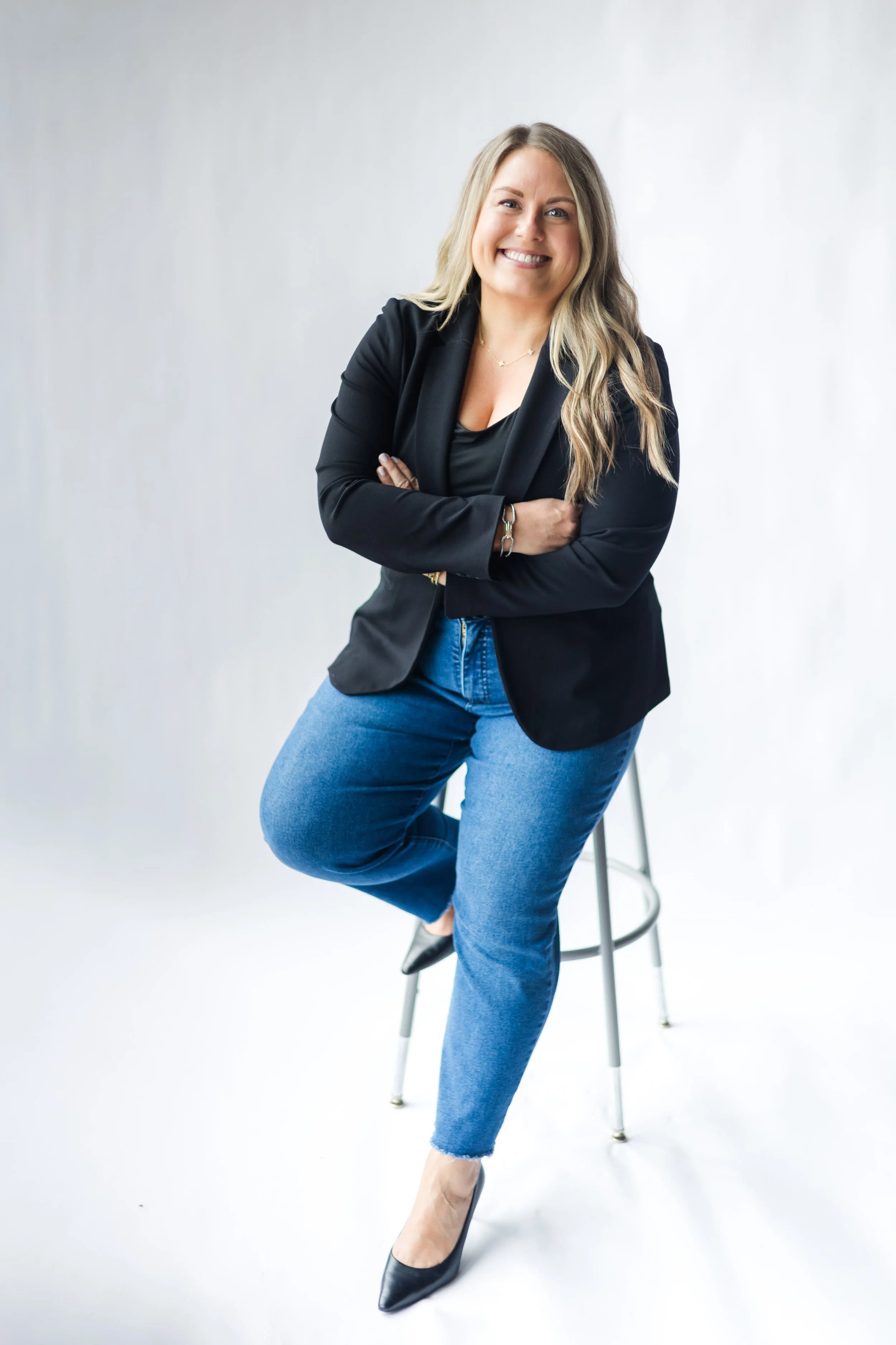 A woman with blonde hair smiling, crossing her arms, wearing a black blazer, black top, blue jeans, and black high heels, sitting on a stool against a white background.