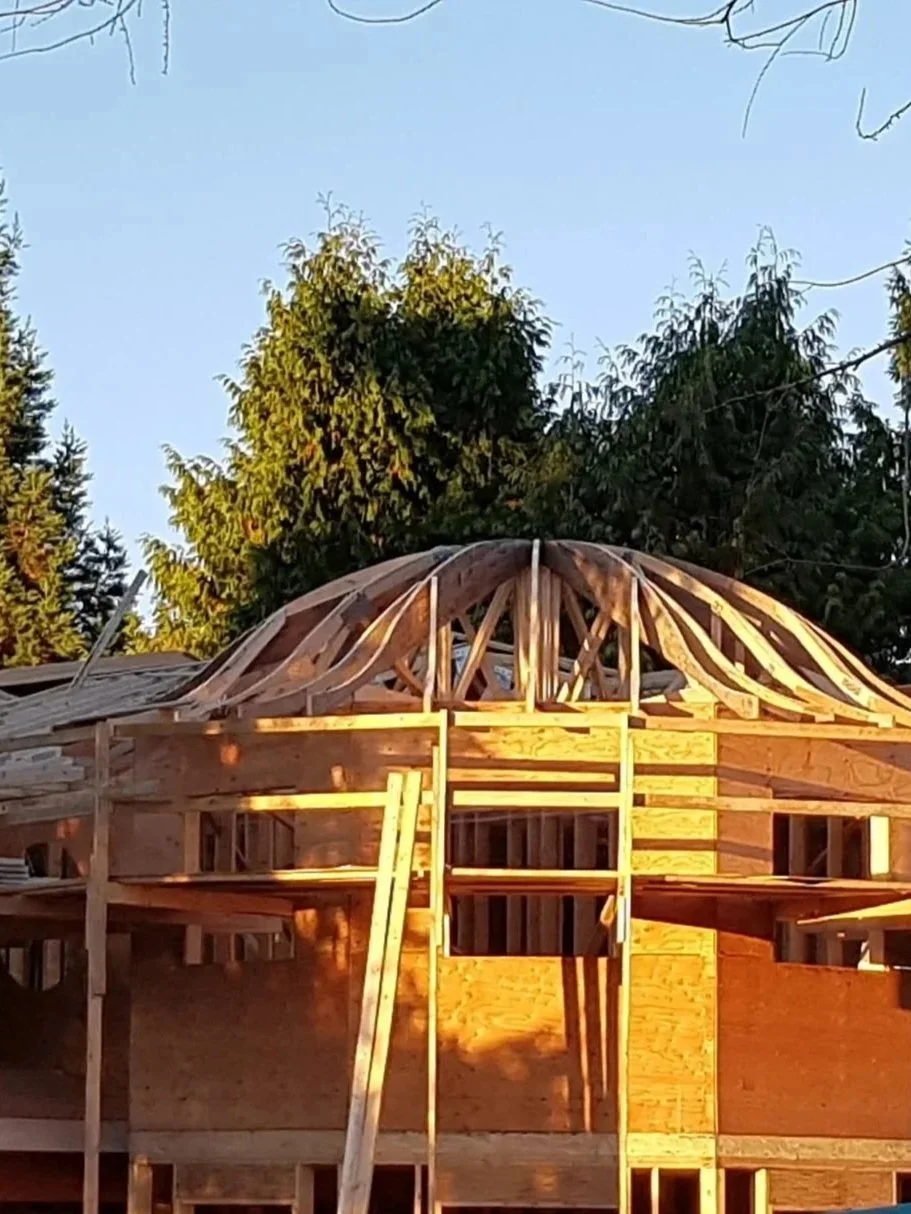 Wooden house under construction in UBC BC with framing and roof support structures in place, surrounded by green trees and a clear blue sky.