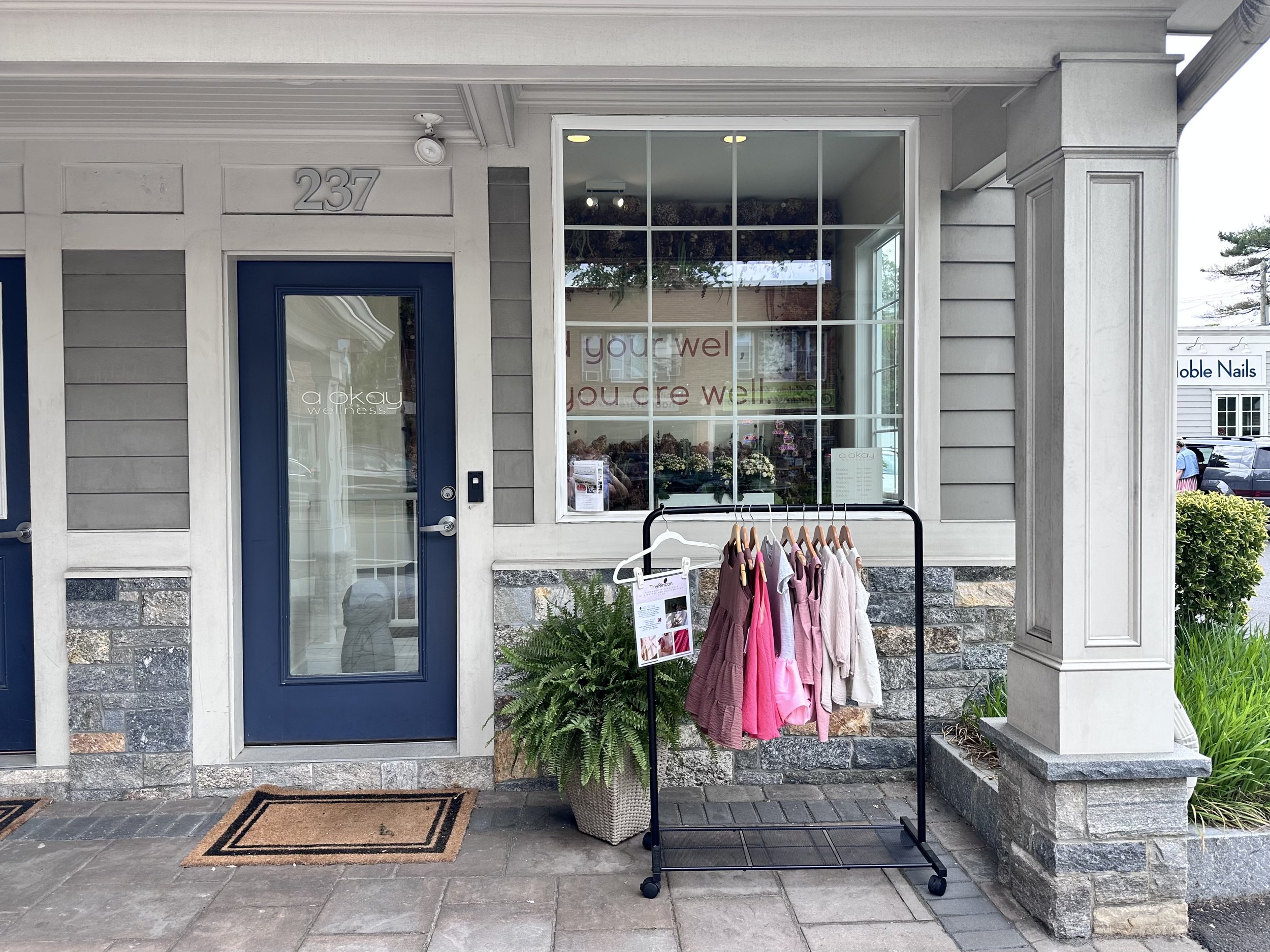 Facade of a building with large window and blue door, with clothing displayed outside on a rack, a potted plant, and signs indicating businesses inside, including Aokay wellness studio, Cos Cob, Greenwich, Connecticut.