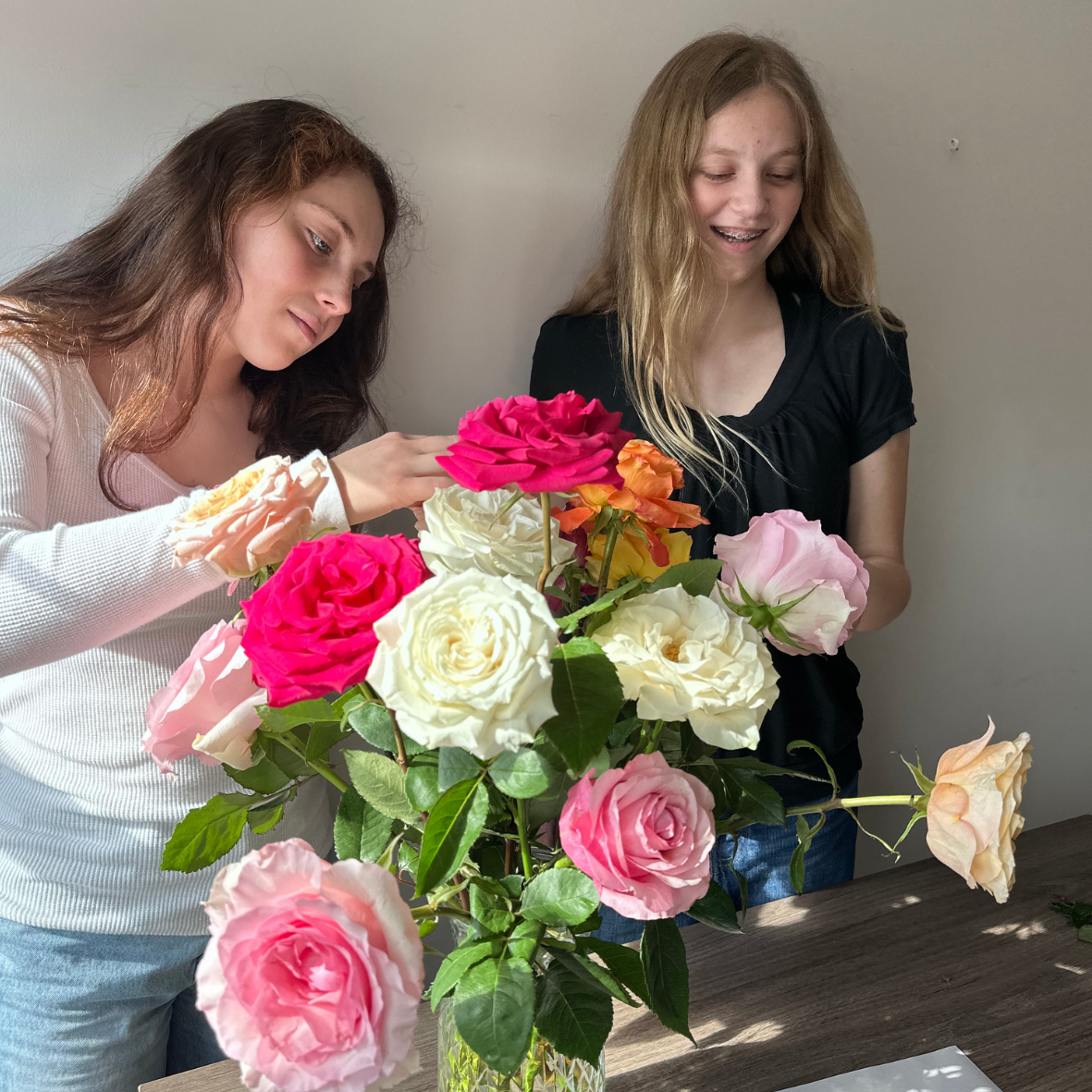 Two young women are standing at a table examining a colorful bouquet of roses in a glass vase.